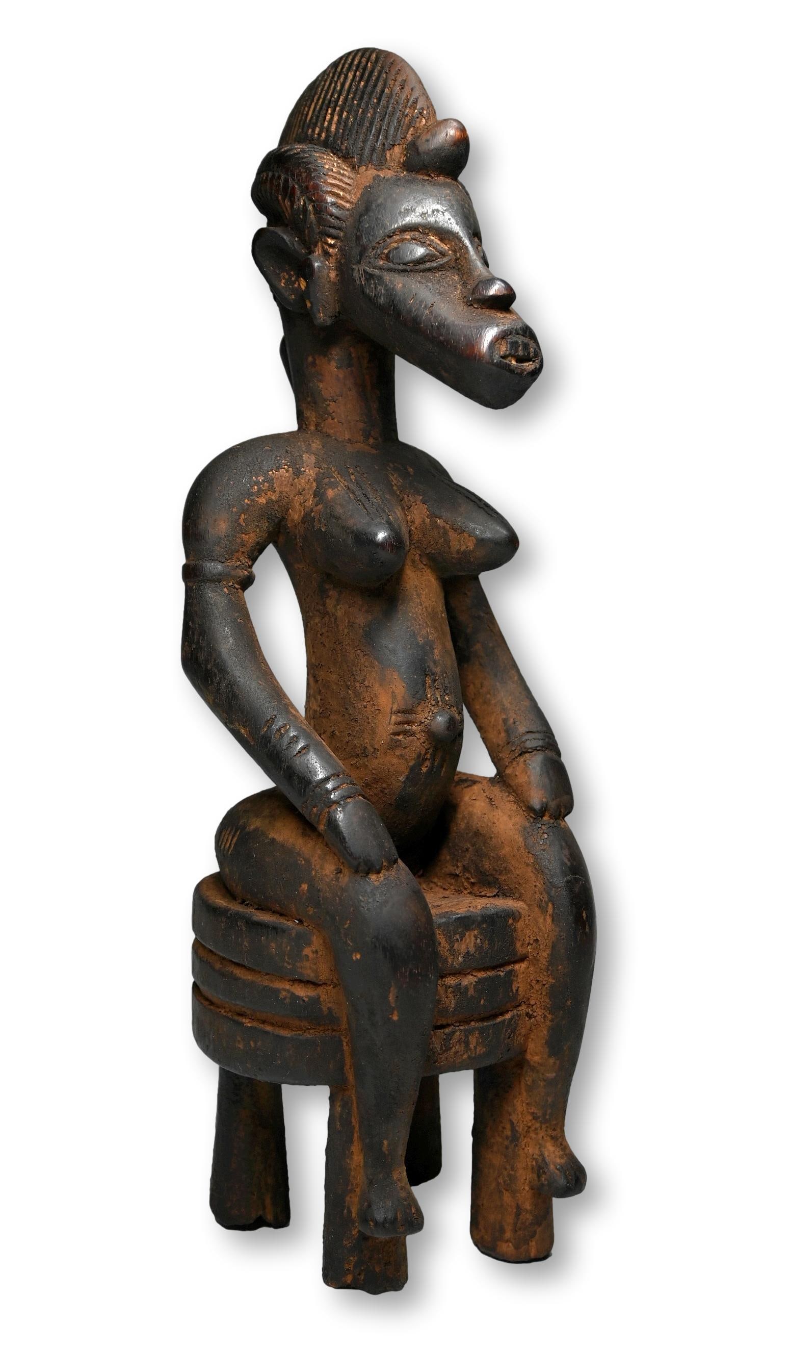 A Senufo Female Sculpture: A Stylized carved wooden female sculpture with elongated features and weathered surface. The Female effigy is shown seated and measures 14.5 inches tall. Senufo peoples, Ivory Coast / Mali, West Afric