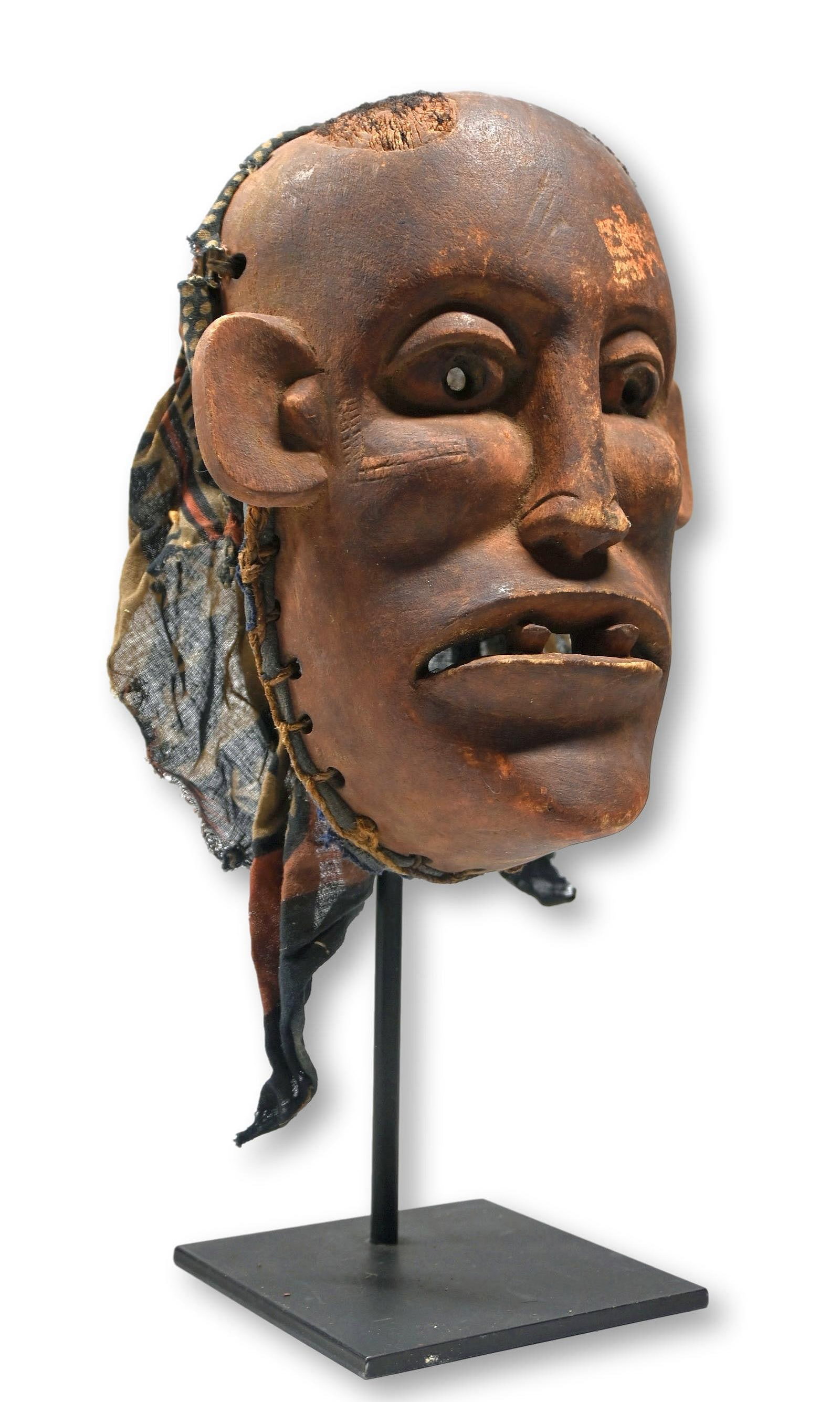 A Makua Ceremonial Mask Field Collected: A Carved wooden ceremonial mask with carved open eyes, raised scar designs, carved filed teeth and two areas of attached human hair on top, embellished with an old batik cloth and painted with red min