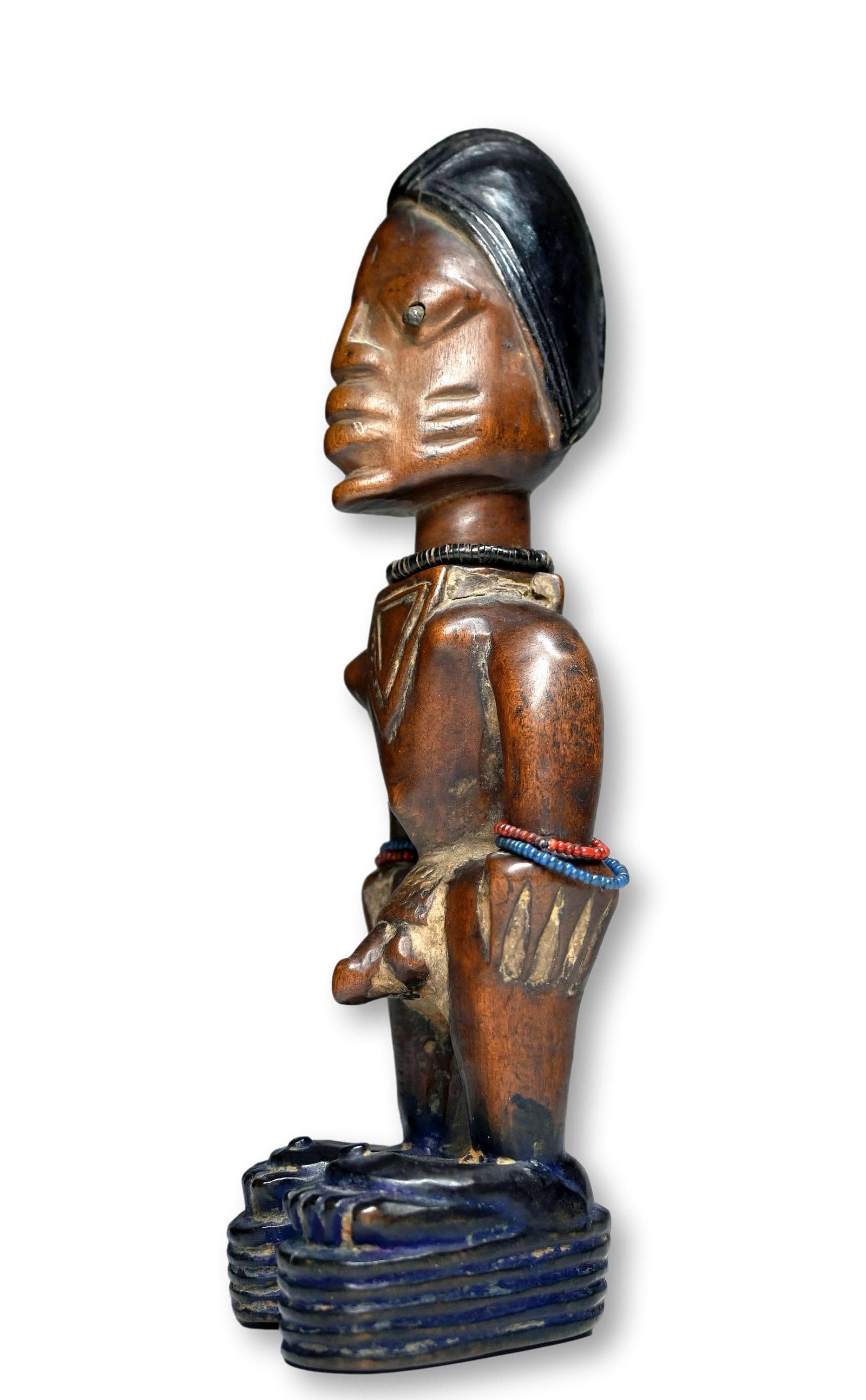 A Yoruba Male Ibeji Ex Steinhacker Gallery 1971: A Classic carved wooden male twin effigy with highly stylized face and tall conical head, the ceremonial cap and feet dyed a deep indigo color, the face worn smooth from ritual feeding. Yoruba Peoples