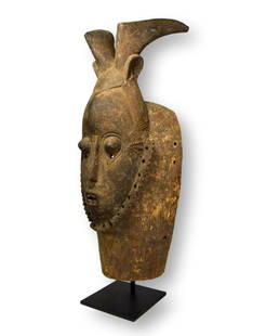 A Baule African mask with Swept Back Horns: A carved wooden mask with raised linear designs, an aggressive face and swept back animal like horns. Baule peoples, Ivory Coast, West Africa. The mask measures 19 inches tall. A metal display stand