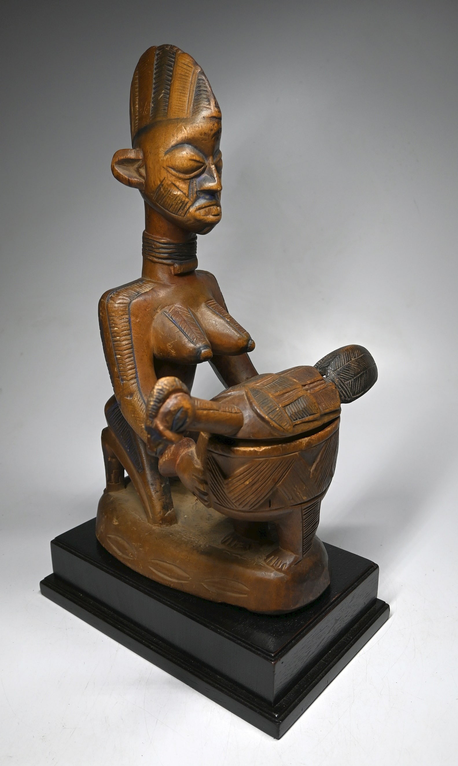A Yoruba Offering Bowl: A finely carved wooden offering bowl known as a Olumeye, which means one who knows respect. The figure of a kneeling woman shown holding and offering a chicken, the top of the bird comes off as a lid 