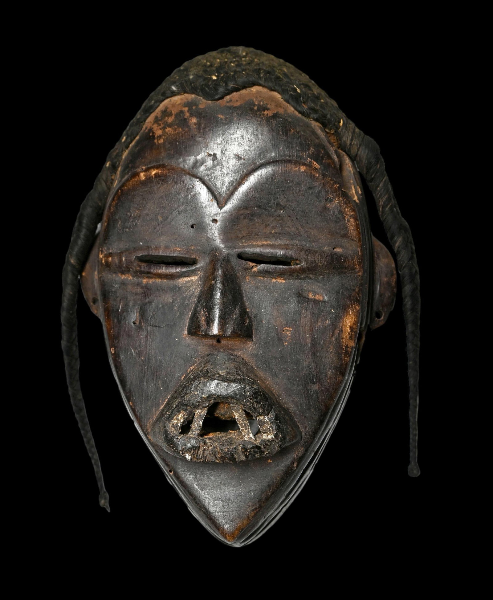 A Dan Mask Ex Bordogna: A Carved wooden mask, likely a deangle mask with attached woven grass fiber coiffure, inset metal teeth , slit eyes and incised linear designs. The mask measures 8.25 x 5 inches. Dan Peoples, Ivory Co