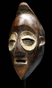 A Mbagani Mask Ex Ravenal Collection: A large carved wood ceremonial mask with characteristic large discoidal eye sockets highlighted in white kaolin with narrow slitted coffee bean shaped eyelids. The domed forehead is capped by a pointe