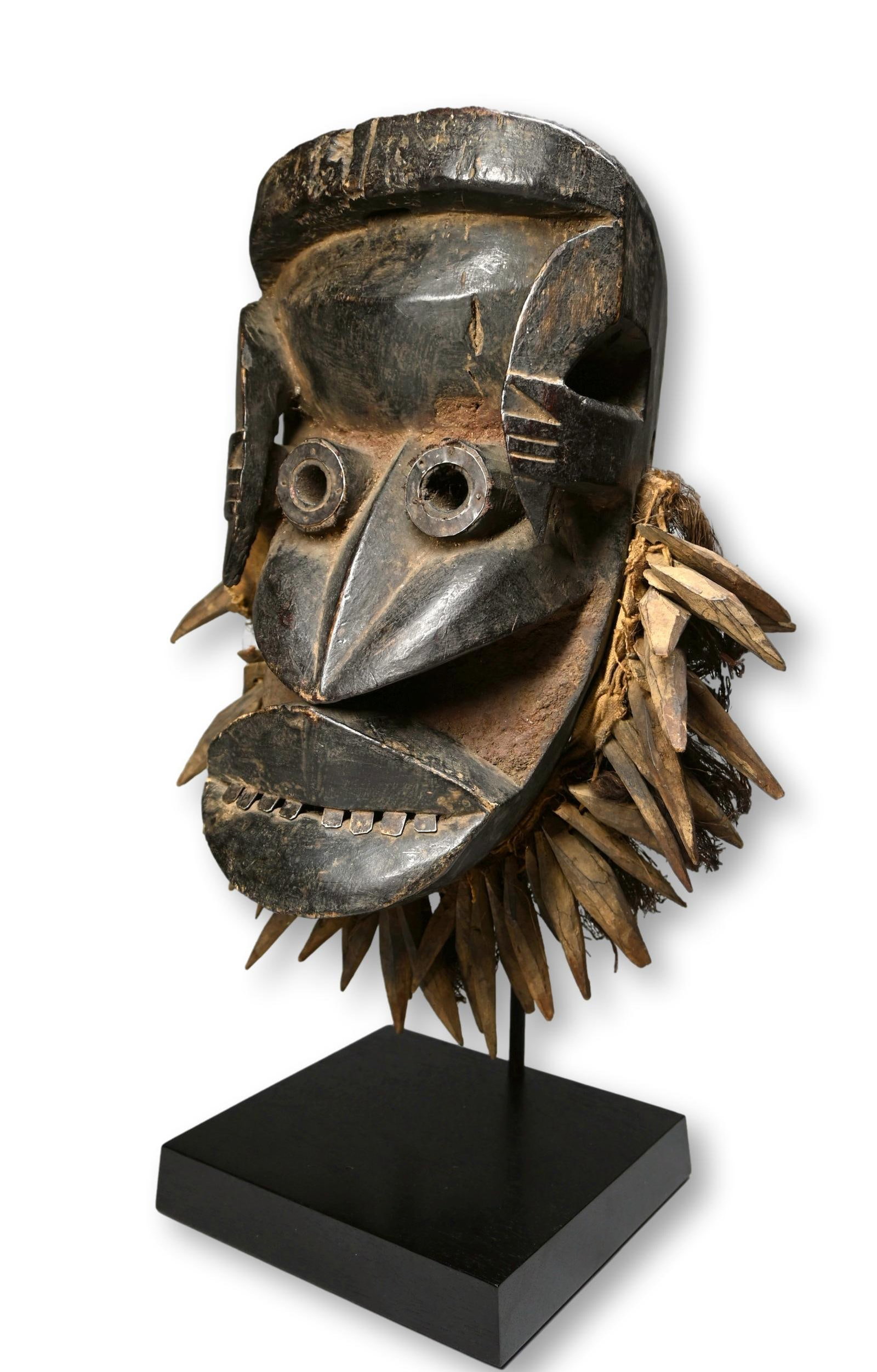 A Dan Guere African Mask: A dark carved wooden mask with inset metal teeth, metal accents around the tubular shaped eyes, stylized hands holding the curved horn like appendages and carved wooden spikes around the perimiter as 