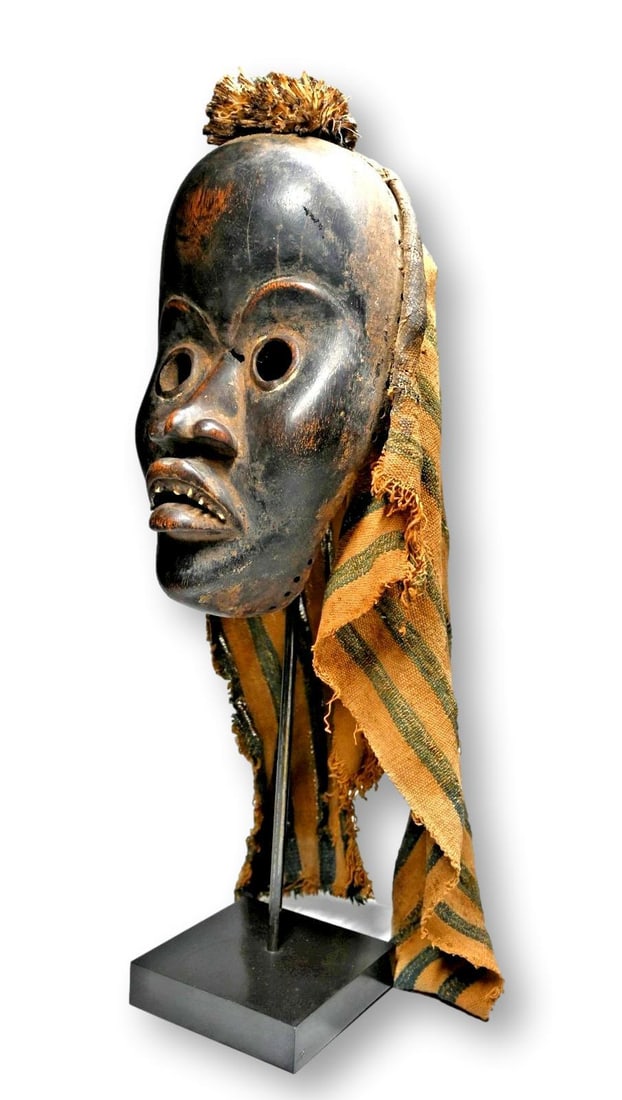 A Dan African Mask: A carved wooden dance mask with open work eyes, realistic facial features including inset teeth , likely animal and a well handled and weathered surface, well worn and painted black.Dan peoples, Liber
