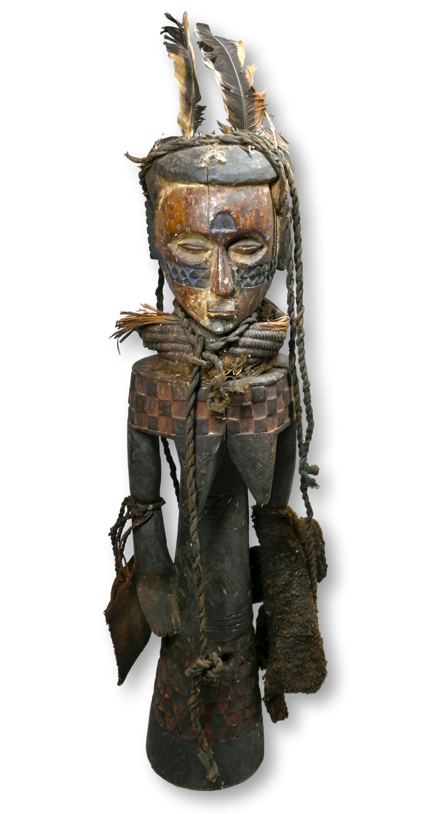 A Kuba Sculpture, African Art: A carved wooden effigy of a ancestor with a mask face and embellished with ropes and feathers. Kuba peoples, Congo, Africa. The sculpture measures 25 inches tall.