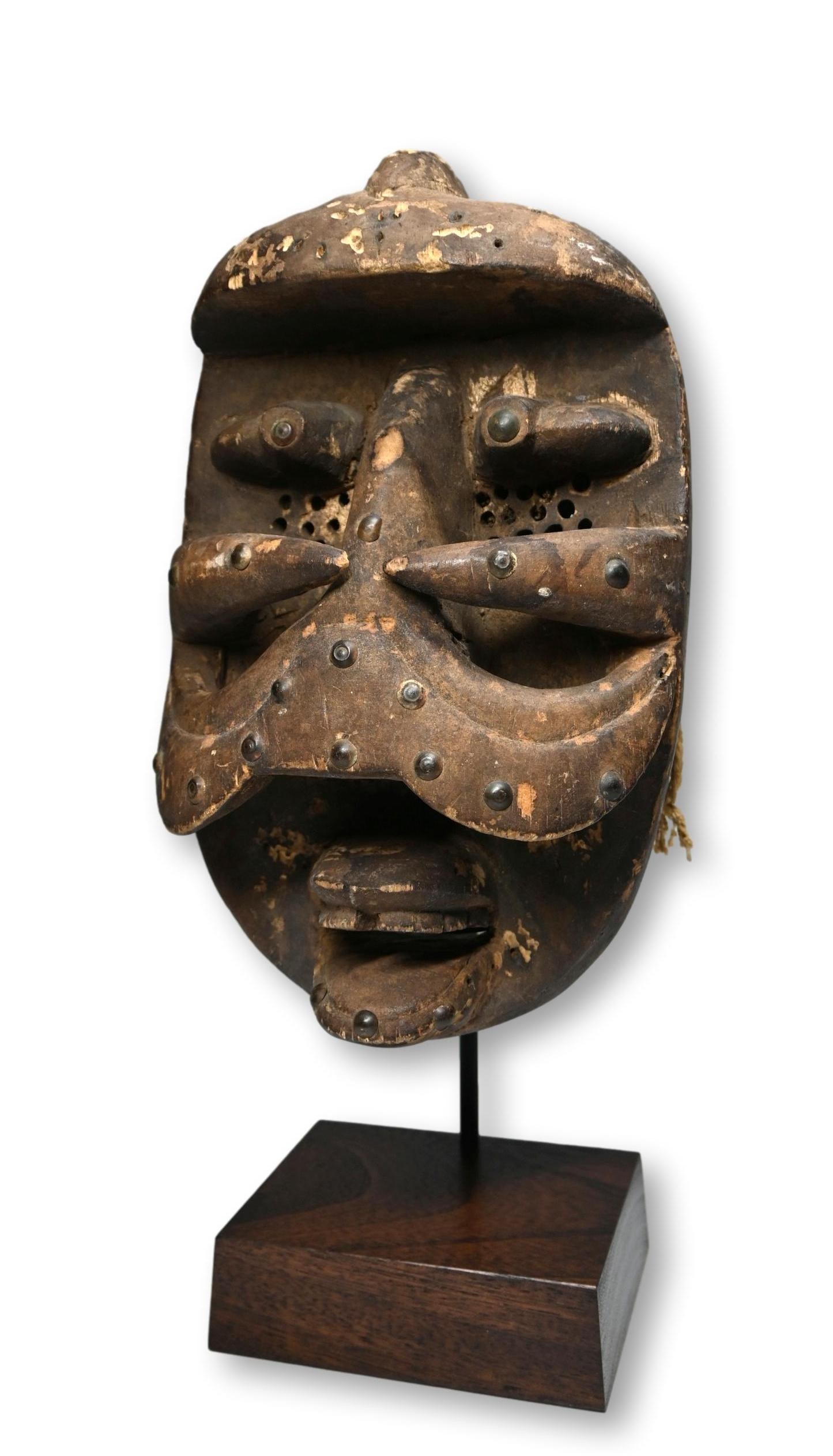 A Fine Old Bete Mask Ex Dannenberg: A carved wooden dance mask with weathered and worn surface, adorned with metal tacks and featuring conical protruding eyes and carved wooden teeth. Bete peoples, Ivory Coast, West Africa, who live nea