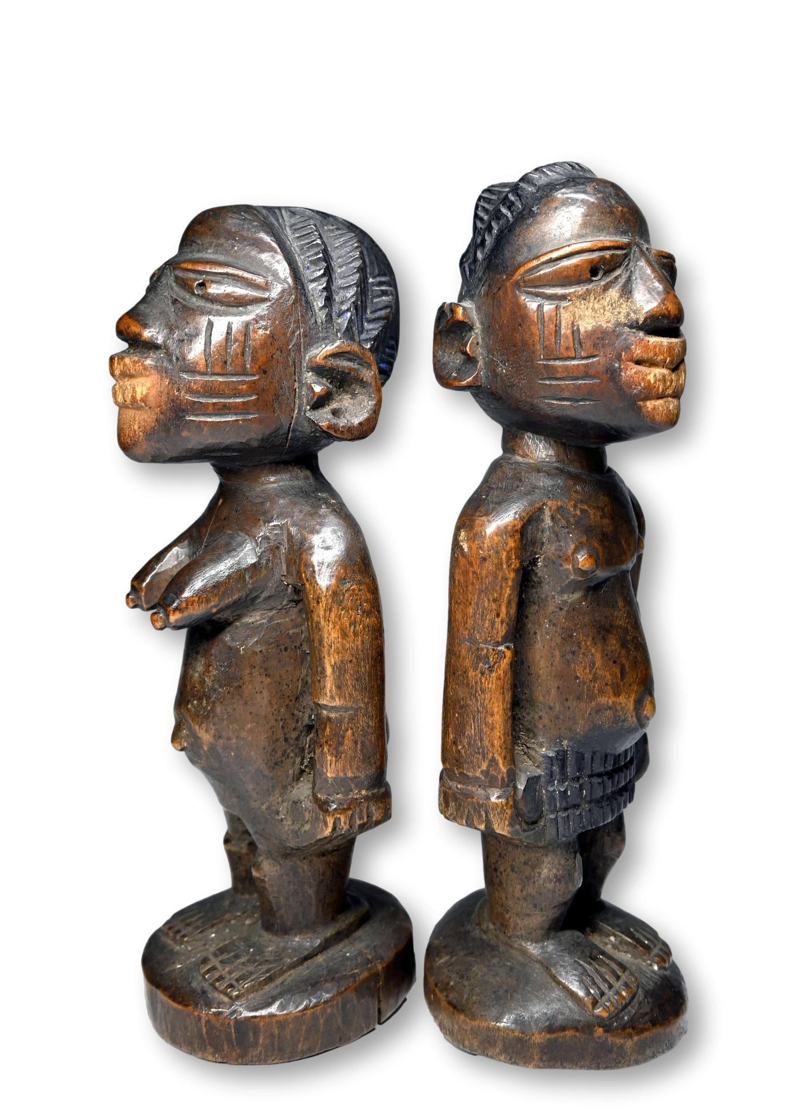 A Pair of Yoruba Ibeji from Igbo Ora: A carved wooden male and female set of twin sculptures with deep facial scars, carved coiffure and a well handled surface patina, note that the sculptures are both well FED, their mouths worn smooth f