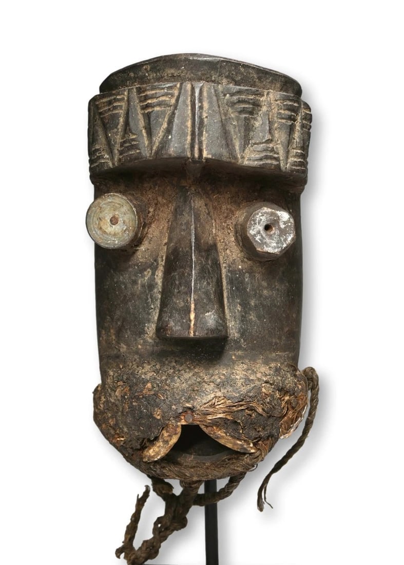 Art Brut Dan Kran Style Mask: A carved wooden mask accented with metal around one of the eyes, large animal tusk teeth and a raised geometric pattern on the forehead that suggests it was carved among the Kran ethnic group within t
