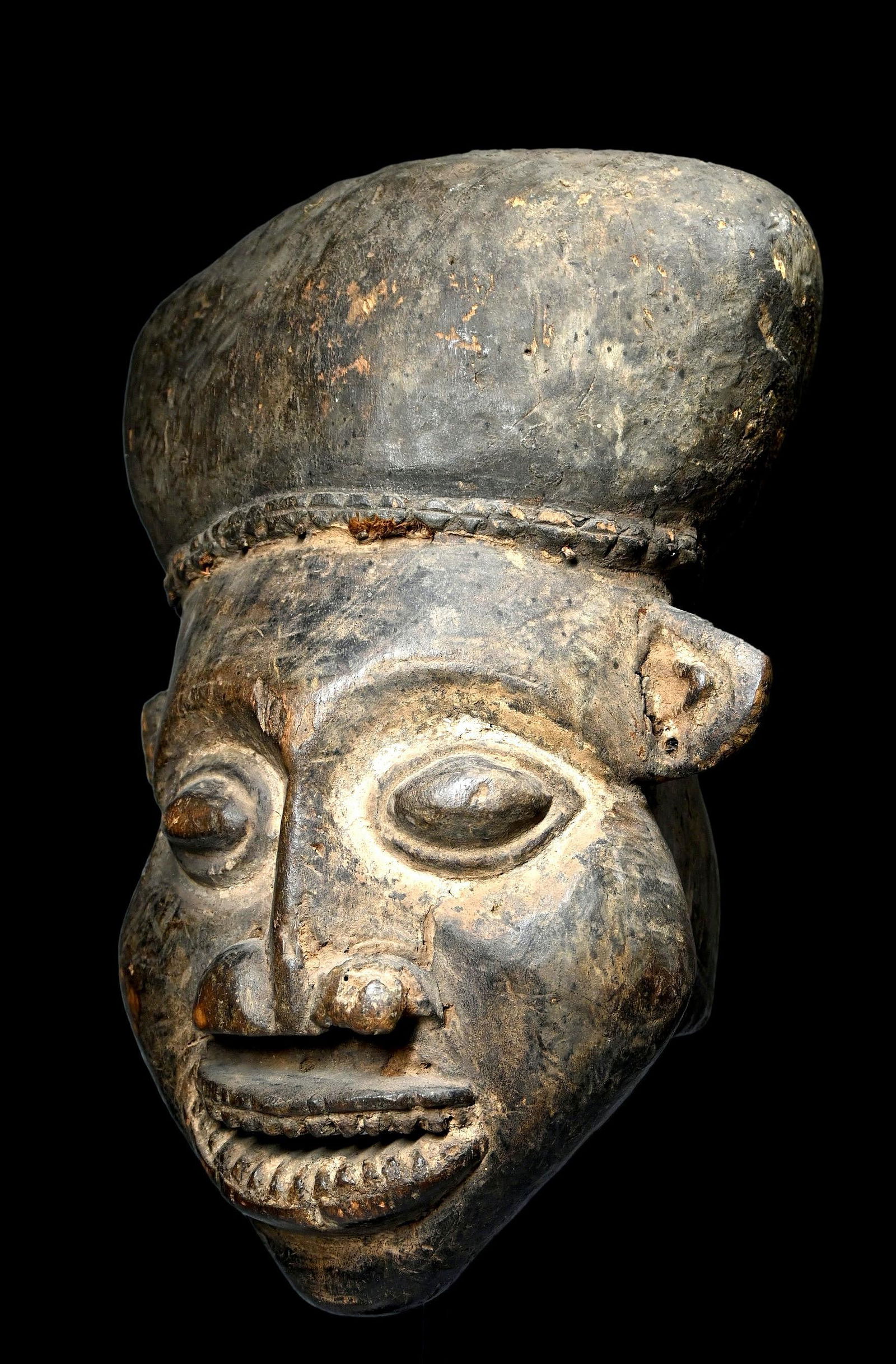 A Kom Helmet Mask: A carved wooden helmet mask with classic facial features, large almond shaped eyes accented with kaolin clay pigments, large smile and layered surface patina as evidence of a long field use as a cerem