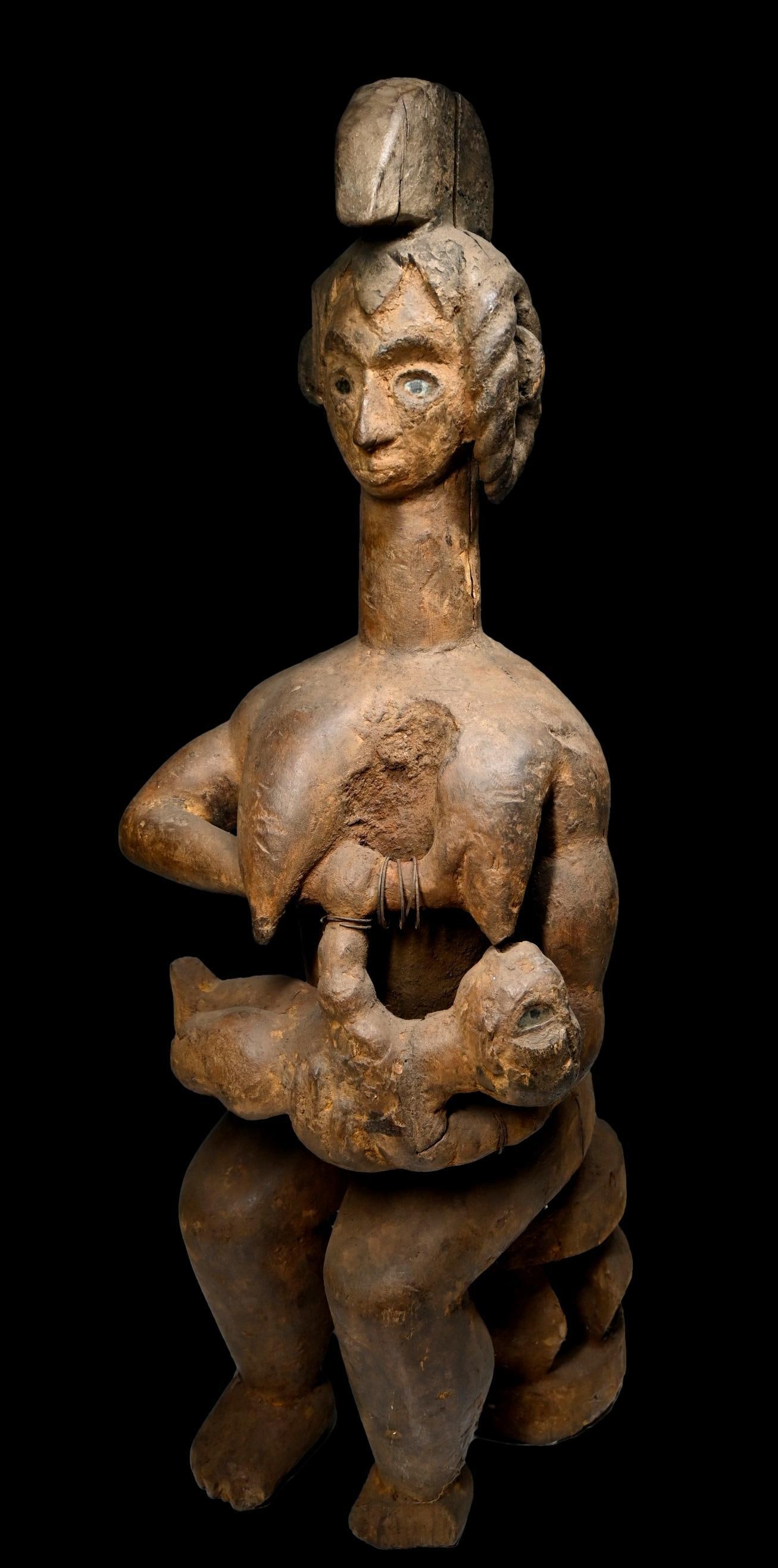A Idoma Maternity Sculpture: A large carved wooden sculpture of a female and child, with inset glass eyes as well as classic treatment to the coiffure and central crest, shown seated. The figure is well weathered and wornm showin