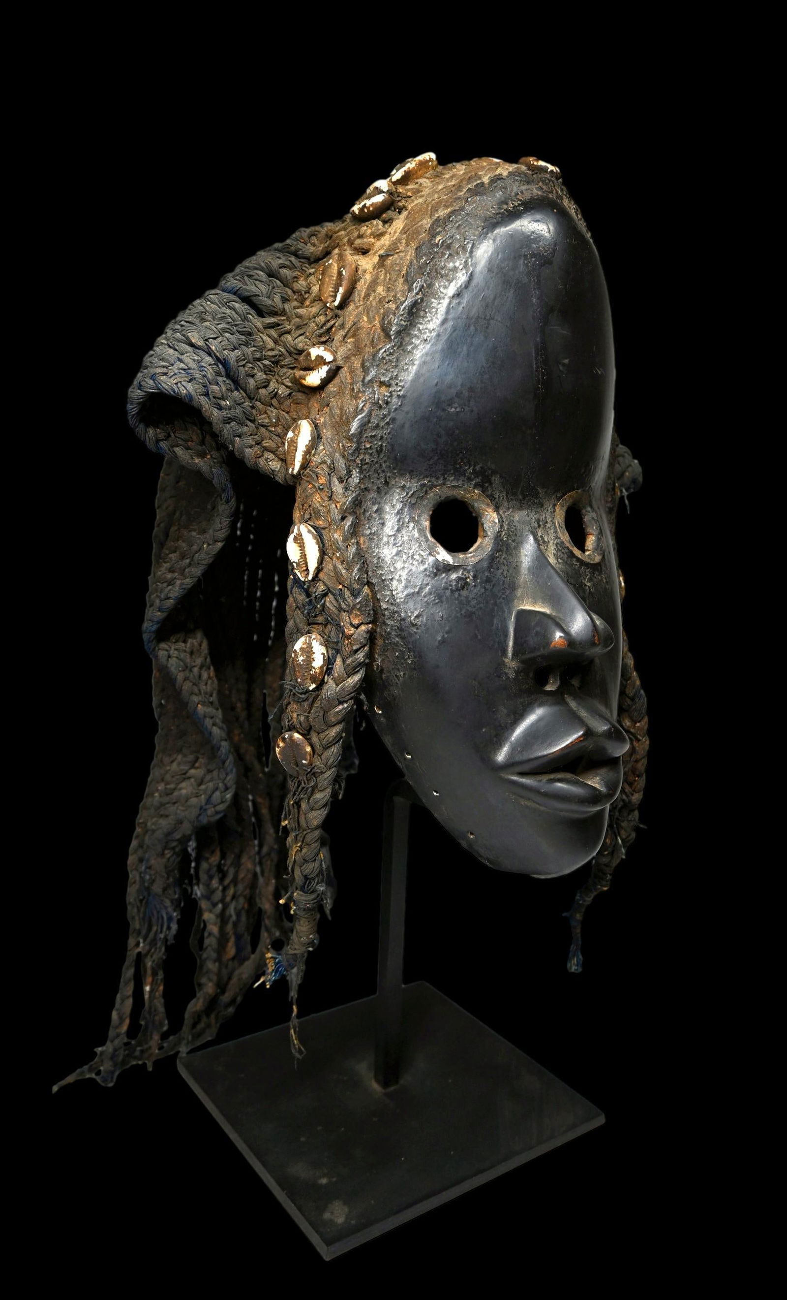 A Dan African Mask: A carved wood and painted dance mask with open work eyes accented with hammered aluminum, woven grass and shell coiffure and woven grass headdress. Dan peoples, Ivory Coast, West Africa. The Mask face