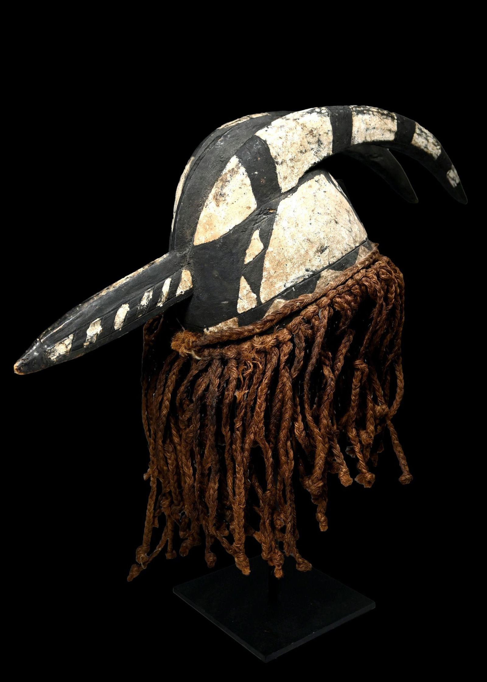 A Mossi African mask: A carved wooden zoomorphic mask with attributes of a bird, and a antelope, and painted with bold contrasting black and white patterns. The bottom of the mask features woven ropes which hang down and c