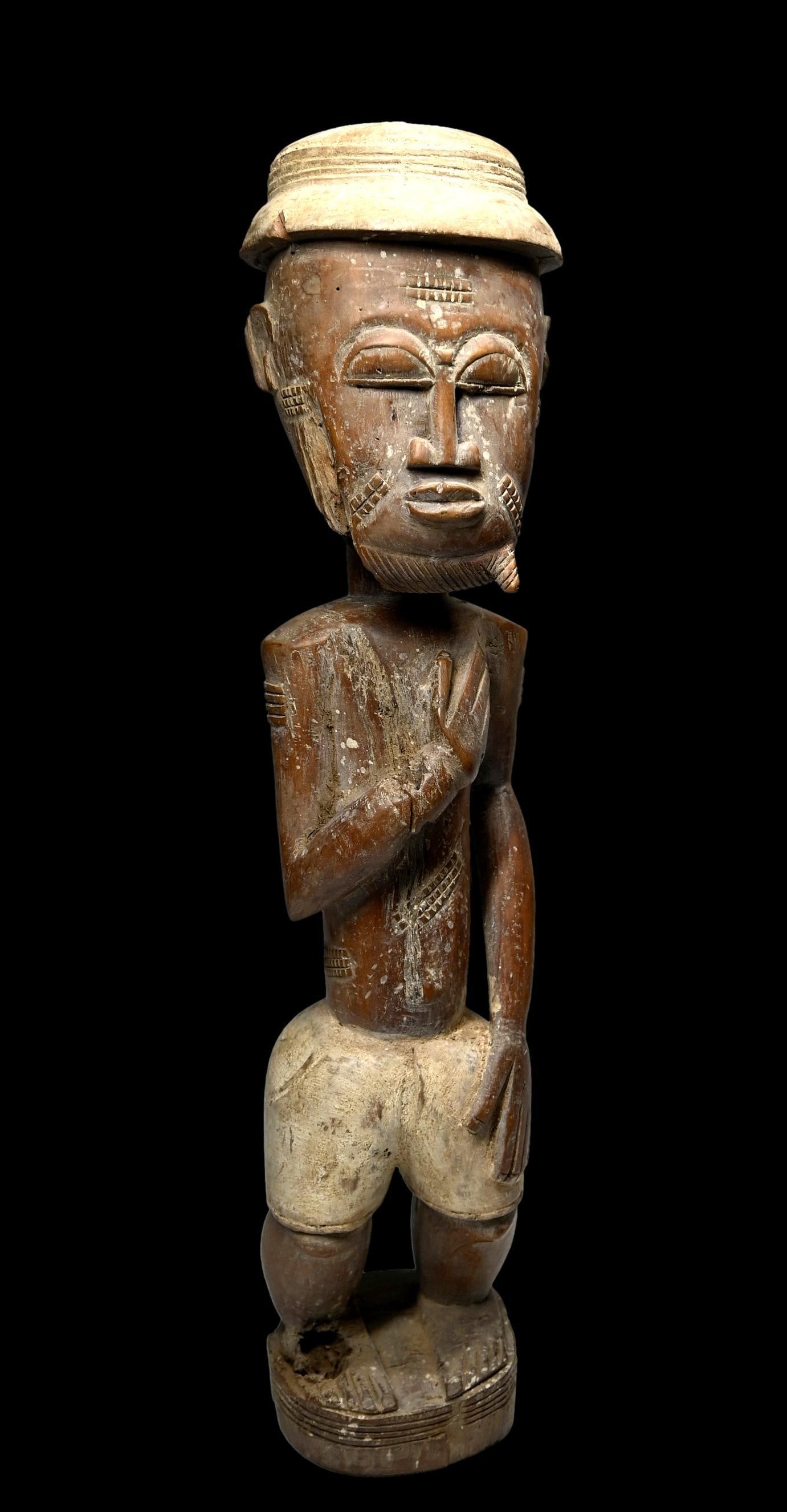 A Baule Colonial Male Sculpture: A carved wood and painted sculpture of a male with classic facial features, scars and beard, wearing a colonial pith helmet and clothing, shown with hands to breast and showing wear and handling. Baul