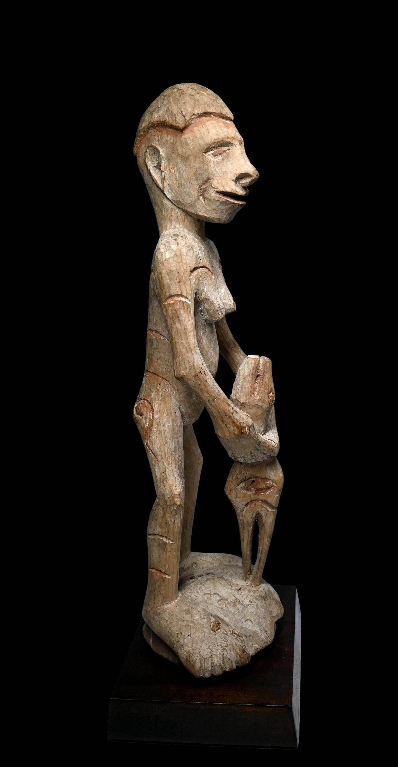 A Female Asmat Ancestor Sculpture: A carved wooden female effigy shown holding a zoomorphic abstract figure, possibly reptile and avian heads, and painted white with kaolin clay pigments and orange or rust colored accents. Asmat people