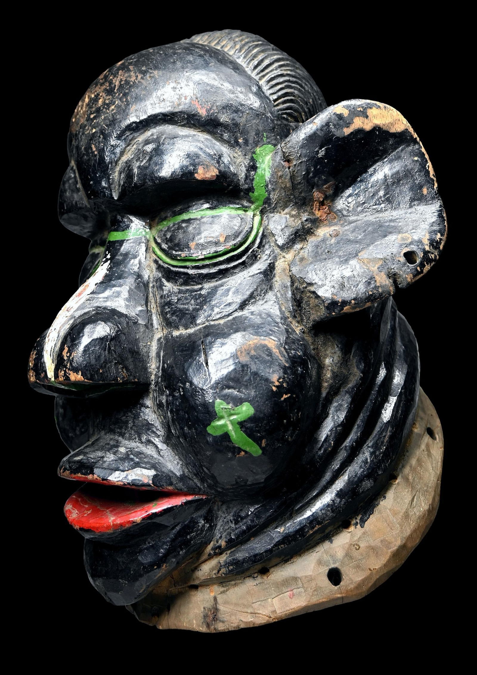 A Ibibio Grotesque Mask: A large carved wooden helmet painted black and embellished with red, white and green designs, including some eye glasses. The mask has exaggerated features like overly large ears and nose, large teeth
