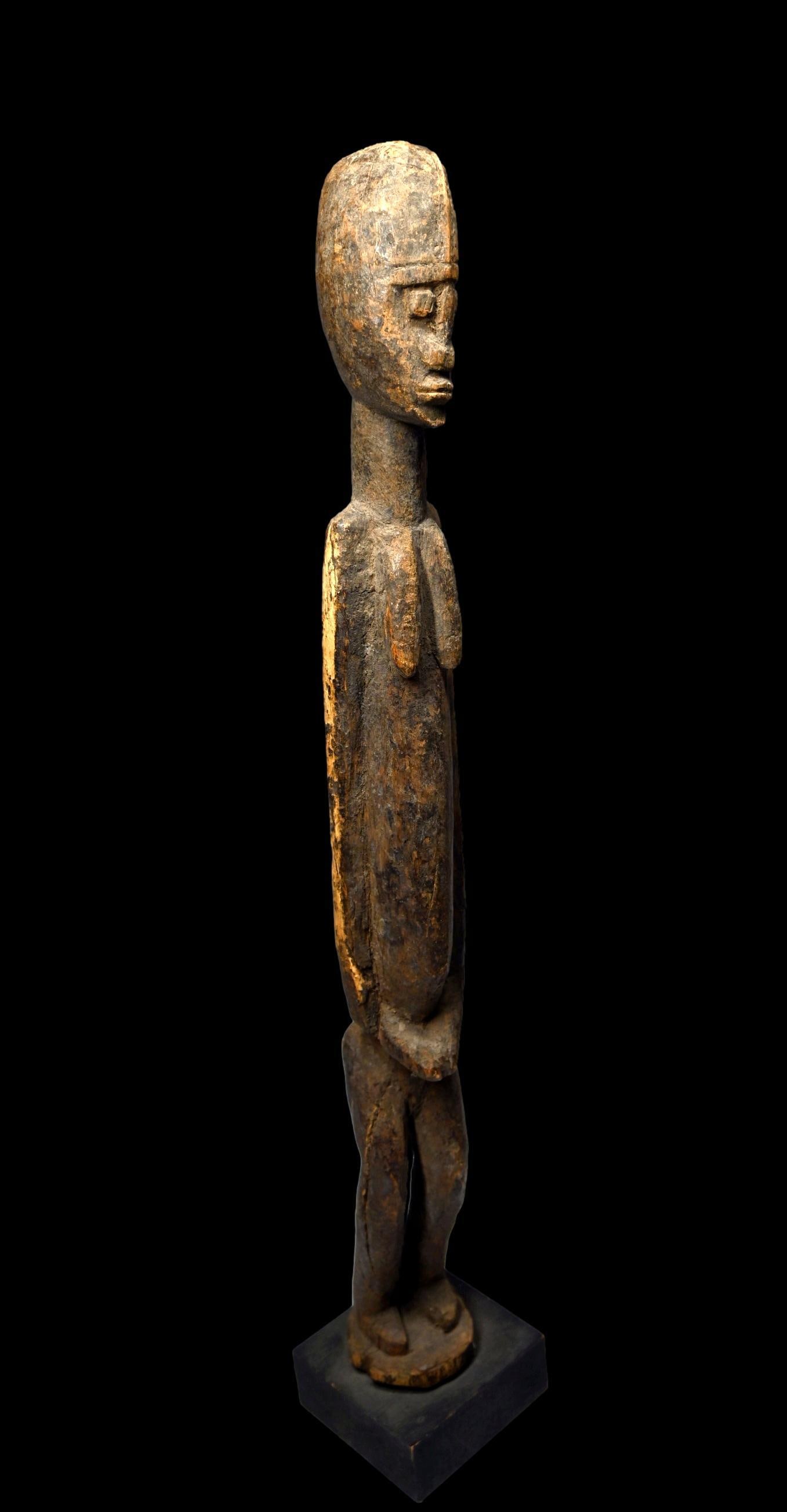 A Dogon Female Effigy: A tall slender carved wooden female effigy with a face and head rendered in a classic style and simple post like form, arms carved tightly to the torso and to the abdomen, the surface suggests some ag