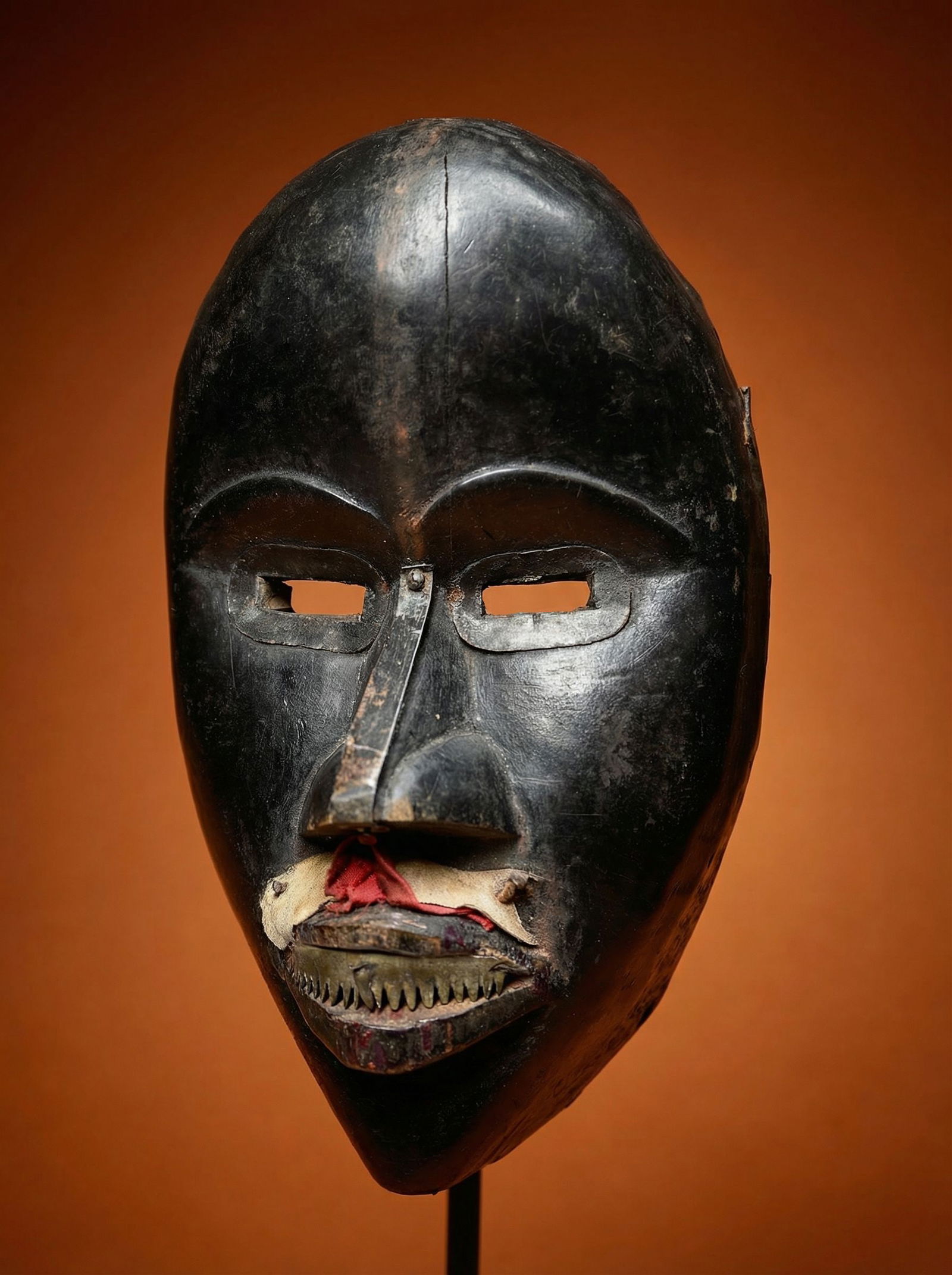 A Dan African mask: A carved wooden ceremonial mask with obvious field use, old nails where textiles and parts of the dance costume where once attached, old metal sharpened teeth and attached metal strips on the bridge o