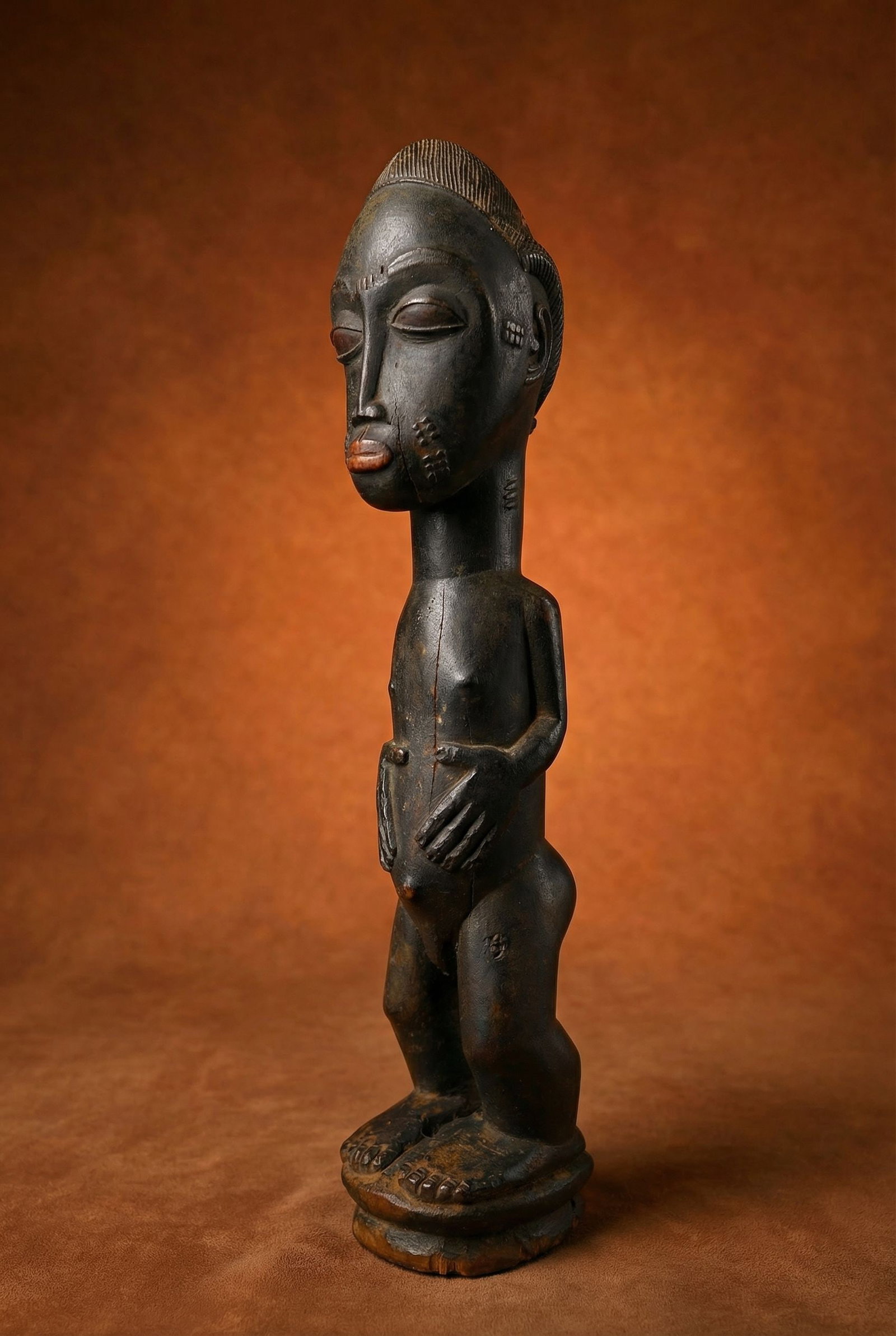 A Baule Male Sculpture: A charming old carved wooden male effigy shown in traditional pose and style, with raised scar designs and elaborate carved coiffure. The sculpture shows plenty of handling and wear and some minor chi