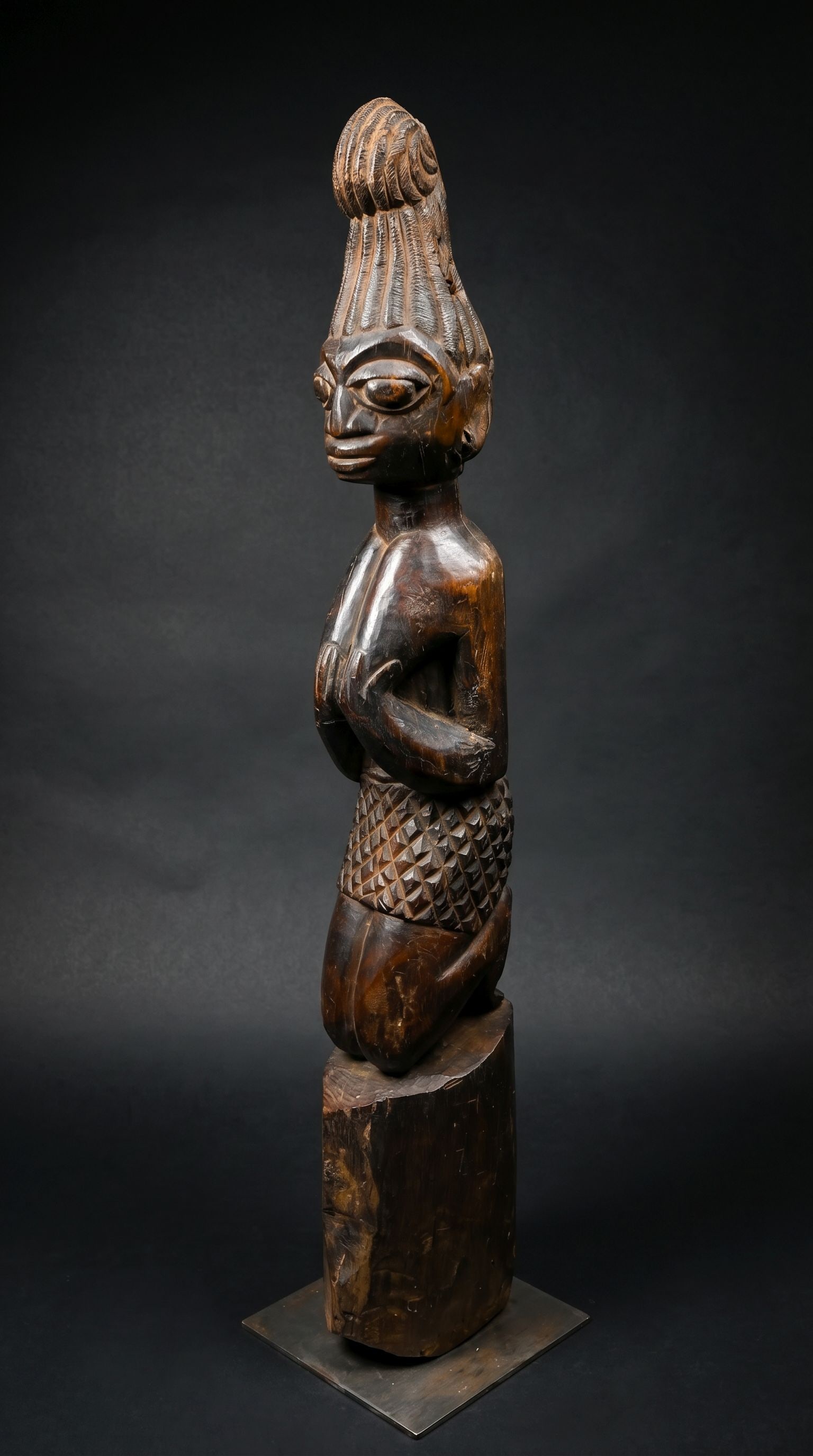 A Yoruba Shango Cult Post with Kneeling Female Devotee: A carved wooden post from a dense and heavy wood featuring the image of a kneeling female figure, her hands to her breast in a sign of fertility and her coiffure set in a elaborate curled crest. Yorub
