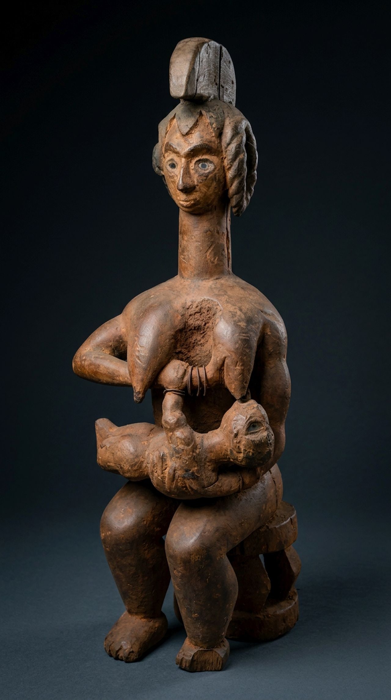 A Idoma Maternity Sculpture: A large carved wooden sculpture of a female and child, with inset glass eyes as well as classic treatment to the coiffure and central crest, shown seated. The figure is well weathered and worn showing
