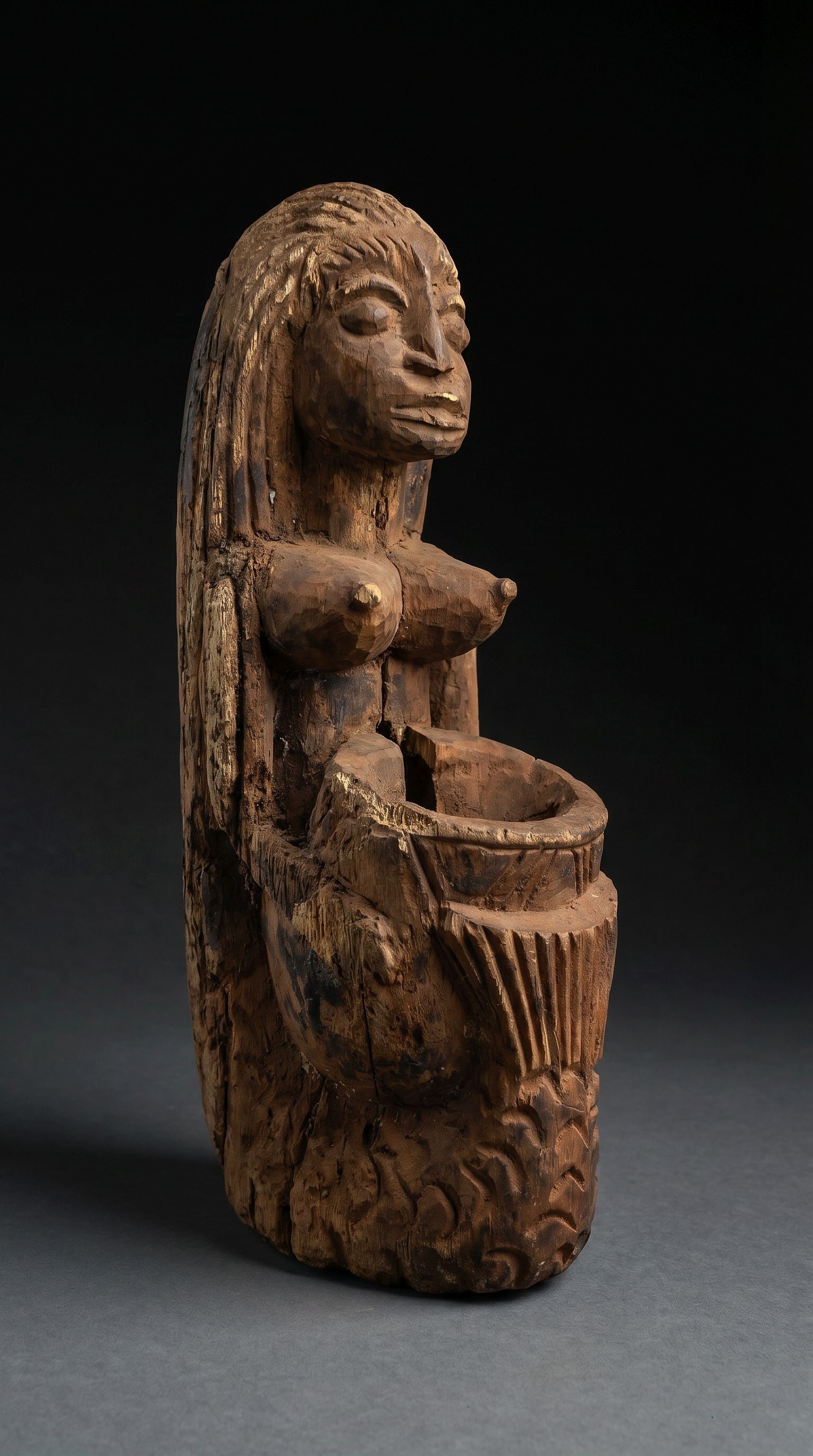 A Yoruba Mami Wata Sculpture: A carved wooden effigy of the Goddess Mami Wata , with mermaid like form, long hair and fish tail, she holds a vessel, presumably for offerings. This is a heavily weathered and eroded example, with