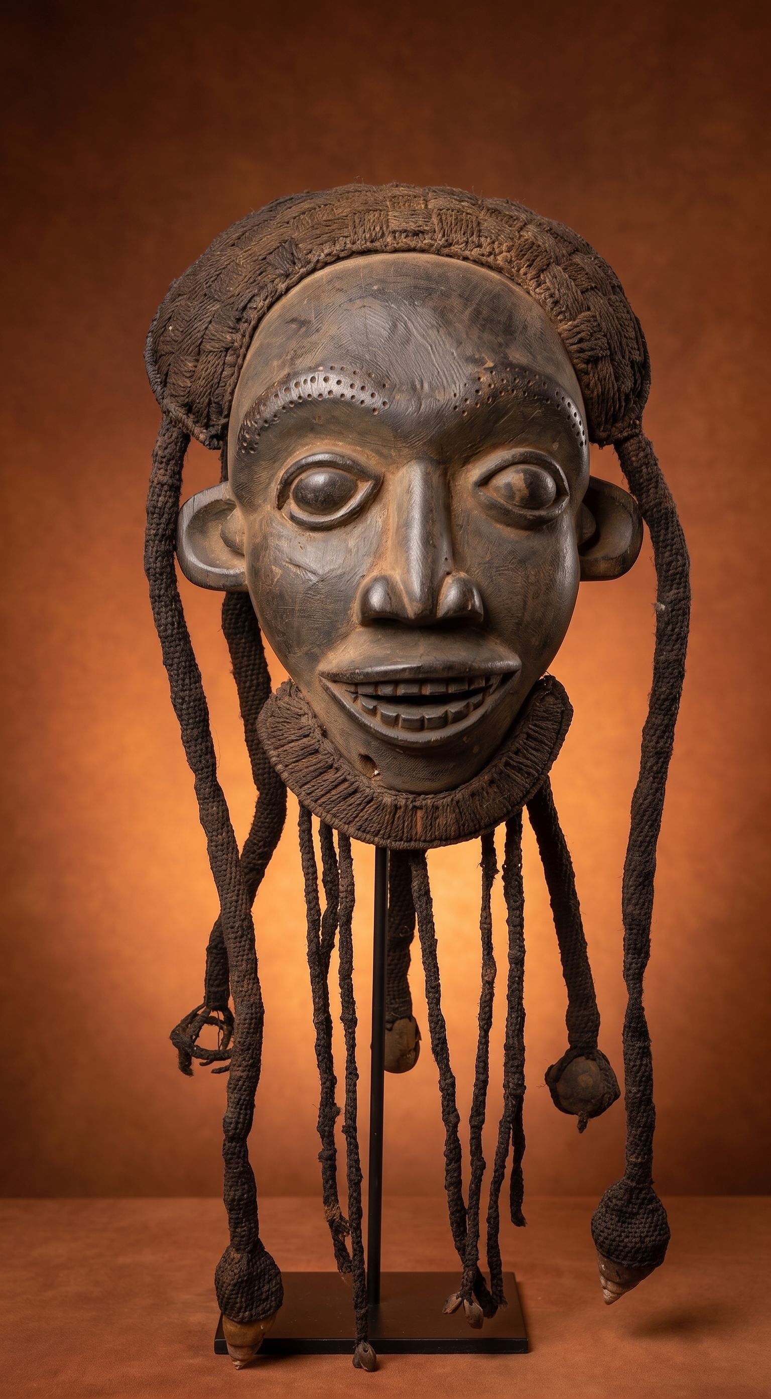 A Bamun Mask with Long Dreadlocks adorned with shells: A carved wooden dance mask embellished with woven plant fiber and long dreadlocks with large sea shells attached as adornment. The mask is well handled and worn and shows some handling and possible fi