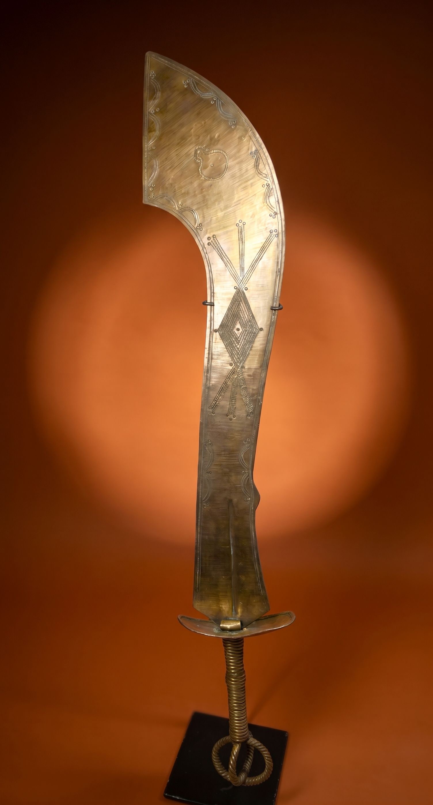 A Brass Ashanti Prestige Sword: A large and elaborate prestige sword with finely crafted blade and twisted metal handle, incised with various tribal motifs. The sword measures 32 x 4.75 inches on a custom display stand. Ashanti peop