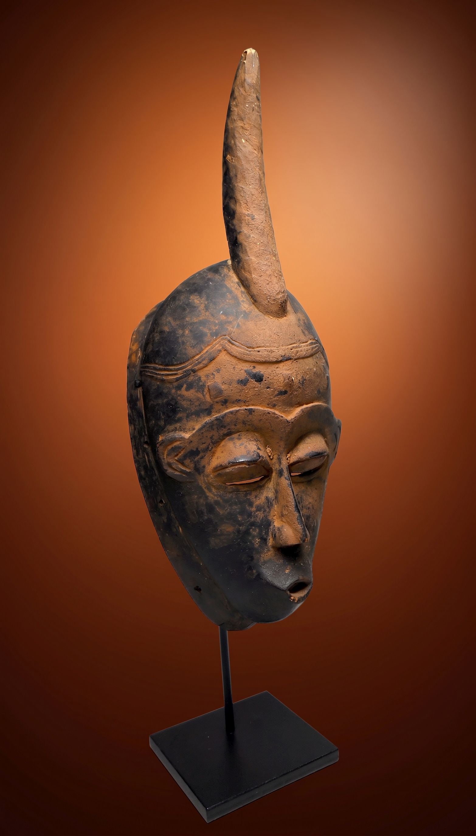 A Serene Baule African Mask: A carved wooden dance mask with a serene face and well weathered surface. Baule peoples, Ivory Coast, West Africa. The mask measures 15 x 6.5 inches , 19 inches tall on custom display stand which is