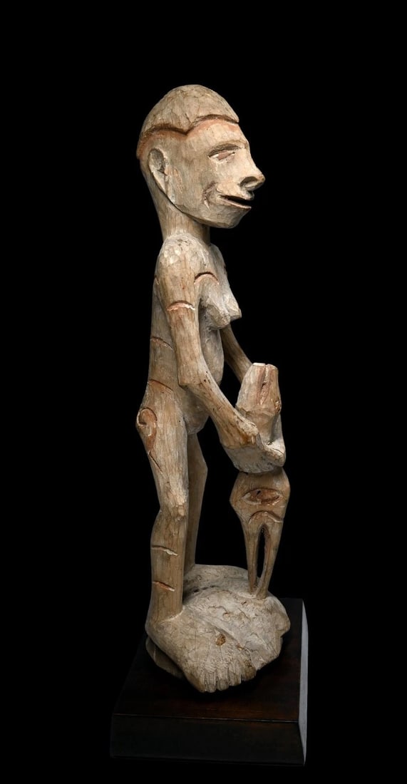 A Asmat Female Ancestor Sculpture: A carved wooden female effigy shown holding a zoomorphic abstract figure, possibly reptile and avian heads, and painted white with kaolin clay pigments and orange or rust colored accents. Asmat people