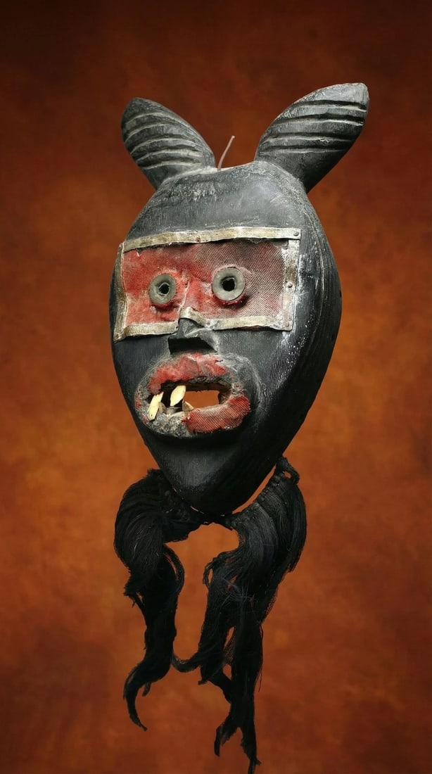 A Dan Mask Collected in the 1960s: A carved wooden mask with stylized horns and accented with aluminum metal strips as well as a red textile and inset real teeth. Dan Mano peoples, Ivory Coast and Liberia, West Africa. The mask measure