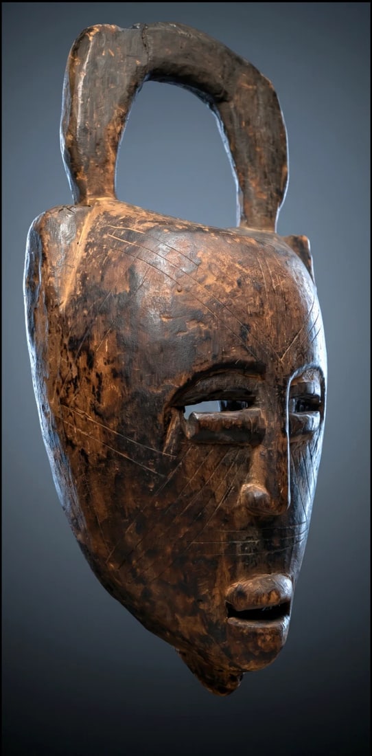 A Bamana African mask: A carved wooden dance mask with a stylized face with incised linear scar like designs and a top arch like finial. The mask showing wear on the interior and obvious weathering on the exterior, restorat