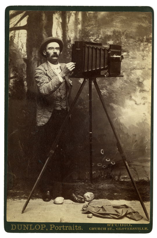 A photographer with a bellows camera, before a: A photographer with a bellows camera, before a landscape backdrop. To increase the illusion there are a few actual rocks on the studio floor. Dunlop, Portraits, Gloversville.