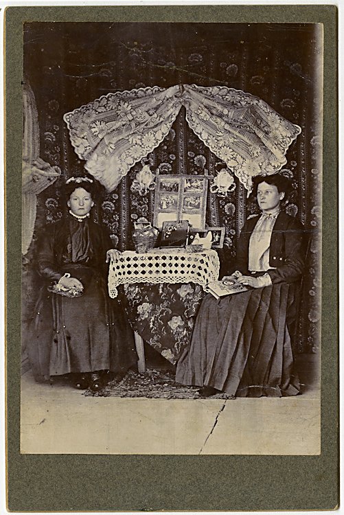 A booth offering cabinet cards and albums.: A booth offering cabinet cards and albums. There’s an upright display of photographs.in back, an album and a basket of photographs on the table. The woman at the right has an album on her lap. The e