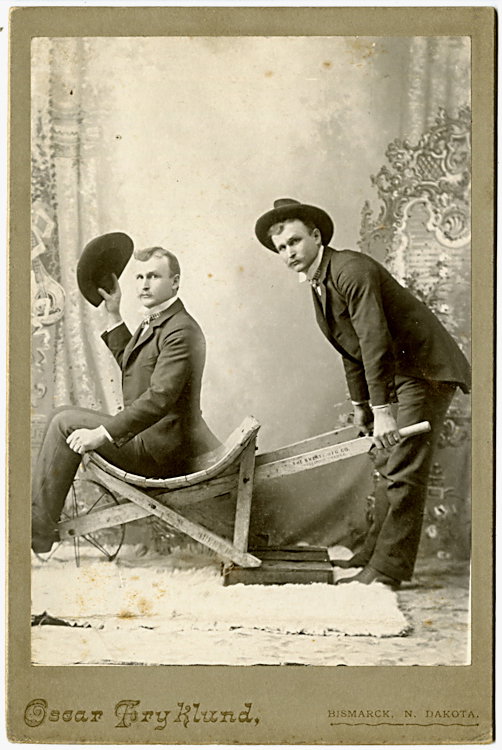 A man pushes himself in a wheelbarrow.: A man pushes himself in a wheelbarrow. The man seated in the barrow lifts his hat. He is the victor in an election, as it was a tradition to push the winner around town in a wheelbarrow. By Oscar Fryk