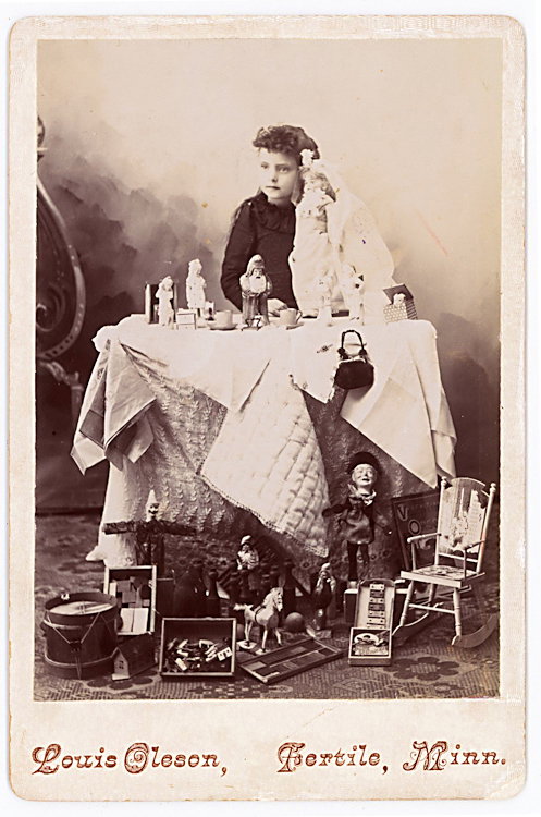 A girl with her doll, toys and other objects on the: A girl with her doll, toys and other objects on the table and floor. They include a puppet, a xylophone, a drum, among other things. By Louis Oleson, Fotile Minnesota.