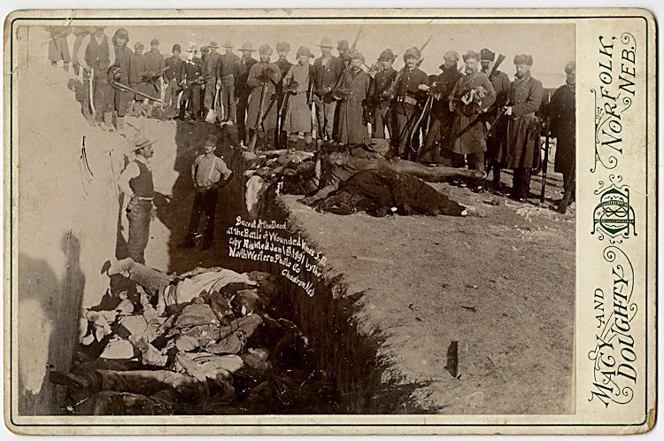 Burying the dead at Wounded Knee.: Burying the dead at Wounded Knee. Workers stand in the mass grave. More corpses lie on the ground ready to be dropped into the grave. Soldiers look on, holding their rifles. One holds a shovel. This i
