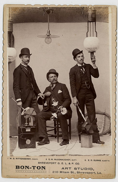 Employees of the Shreveport G. E. L. & P. Company.: Employees of the Shreveport G. E. L. & P. Company. The engineer is at left. A fan is on top of the meter in front of him. In the center sits the Superintendent, holding a ceiling fixture. At right is 
