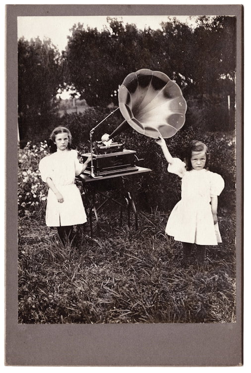 A large cylinder gramophone with a large speaker.: A large cylinder gramophone with a large speaker. Girls pose with it outside. This is likely a prize possession of their father’s that was brought outside to be photographed. No maker indicated.