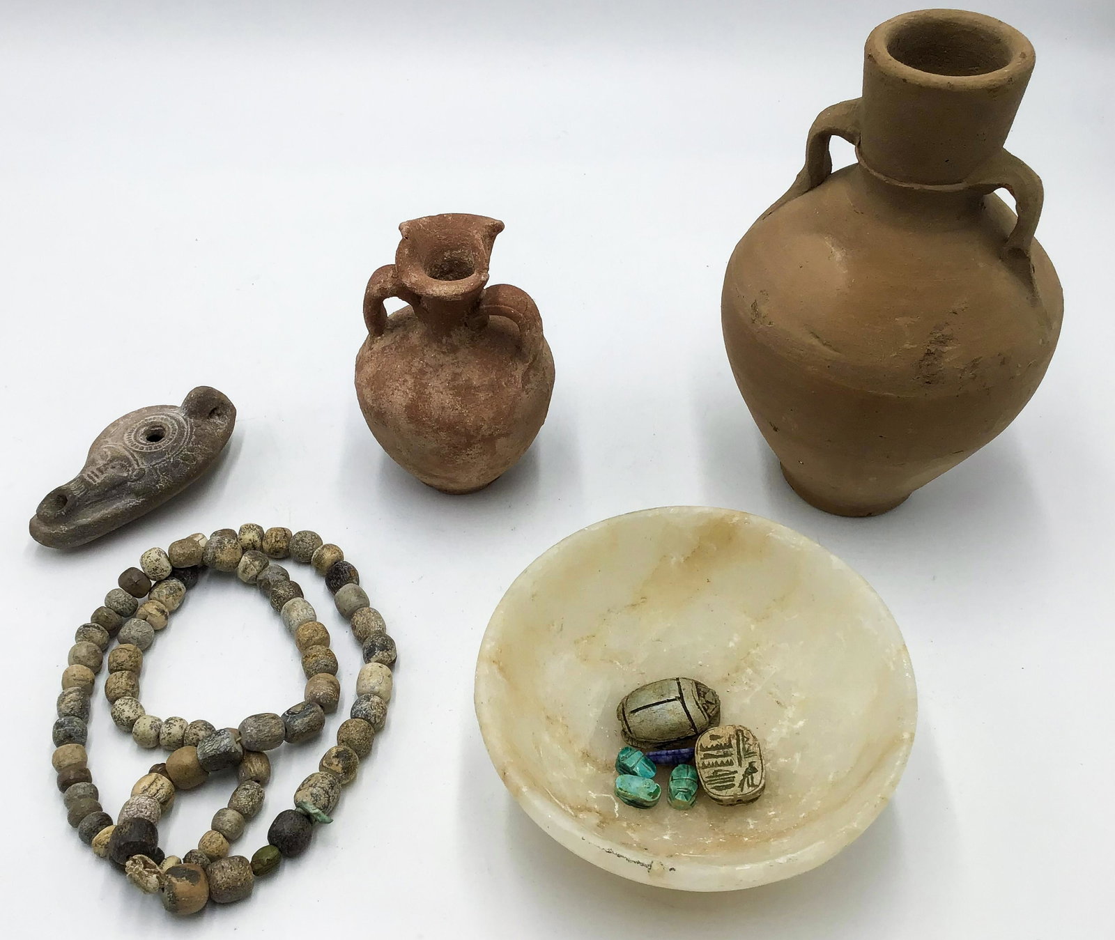 Lot of 11 Egyptian Articles.: Includes: One alabaster bowl, Two Clay Vases, One Clay Oil Lamp, a Carved Stone Bead Necklace containing Two Amulets and Two Scarabs, and Six Separate Scarabs. Third-Party Shipping Required.