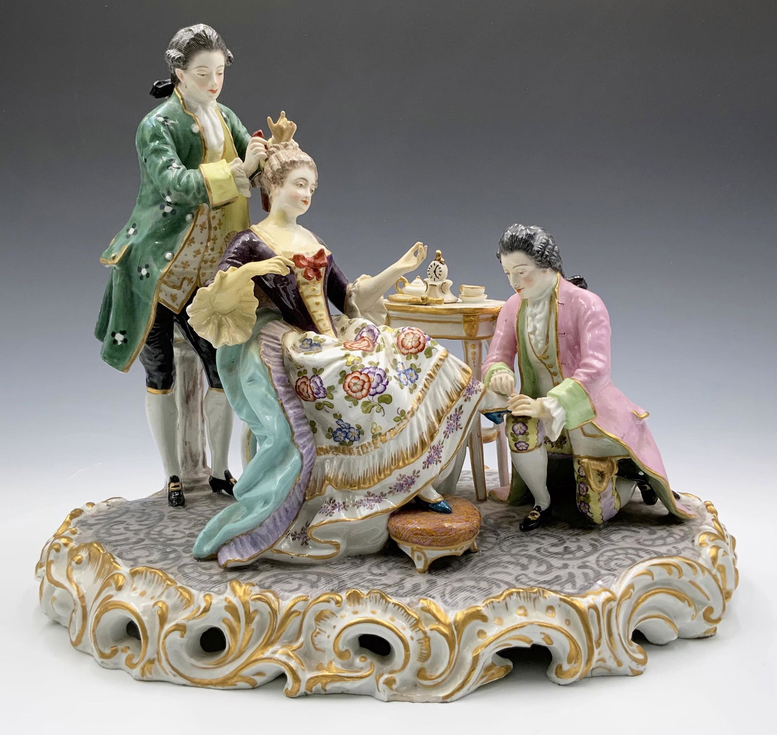 Meissen Figural Group of Woman Being Preened.: A hand painted figure, with gold finish along the bottom. The mark by the white column features a pair of blue double crossed swords. One man is putting a decoration in the lady's hair, the other is a