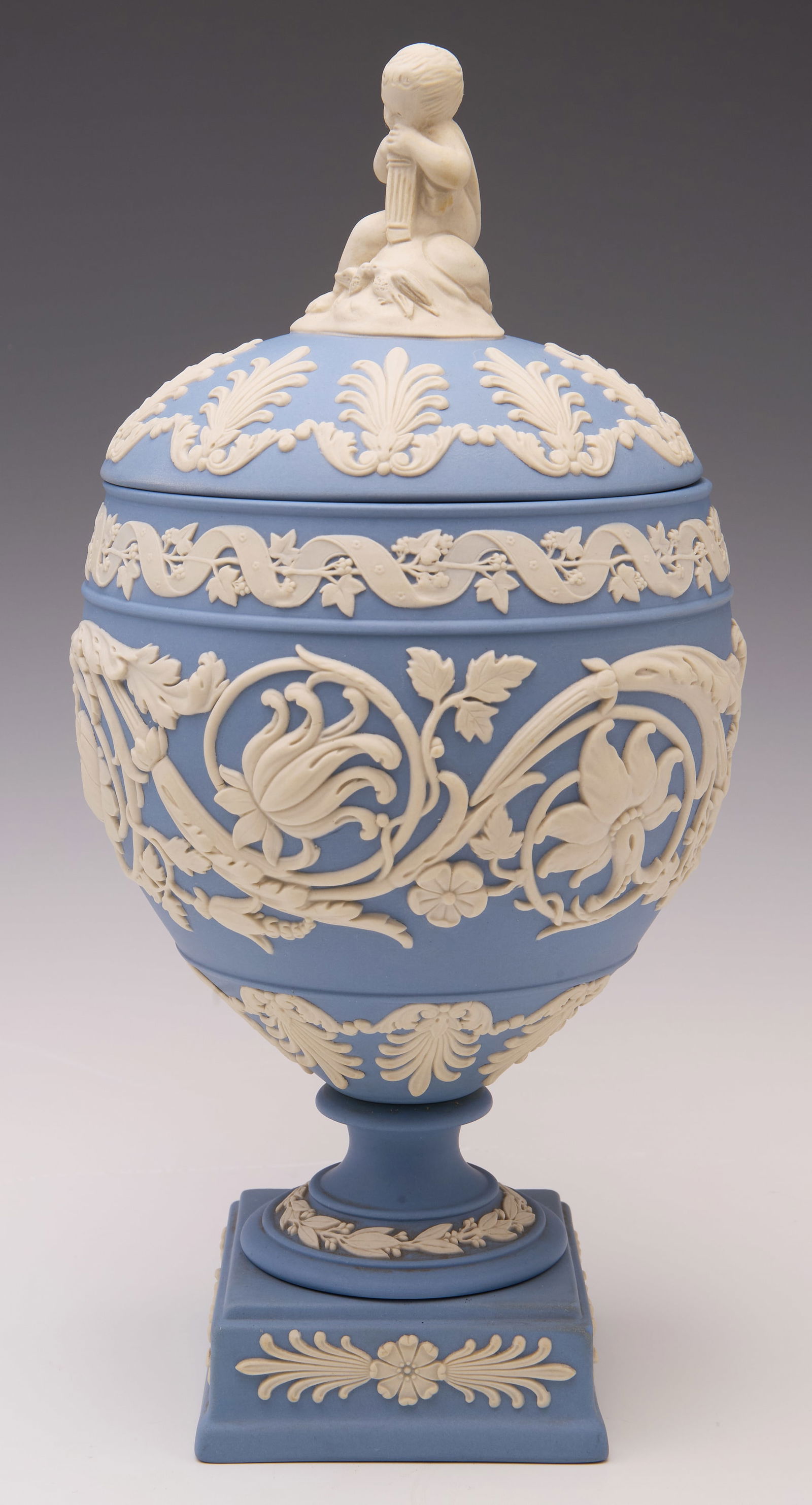 Wedgwood Jasperware Blue & White Arabesque Urn.: The underside is stamped, "Wedgwood Made in England". The lidded urn is blue and white, decorated with elaborate flowers. The finial resembles a small child with two doves. Overall, this piece looks t