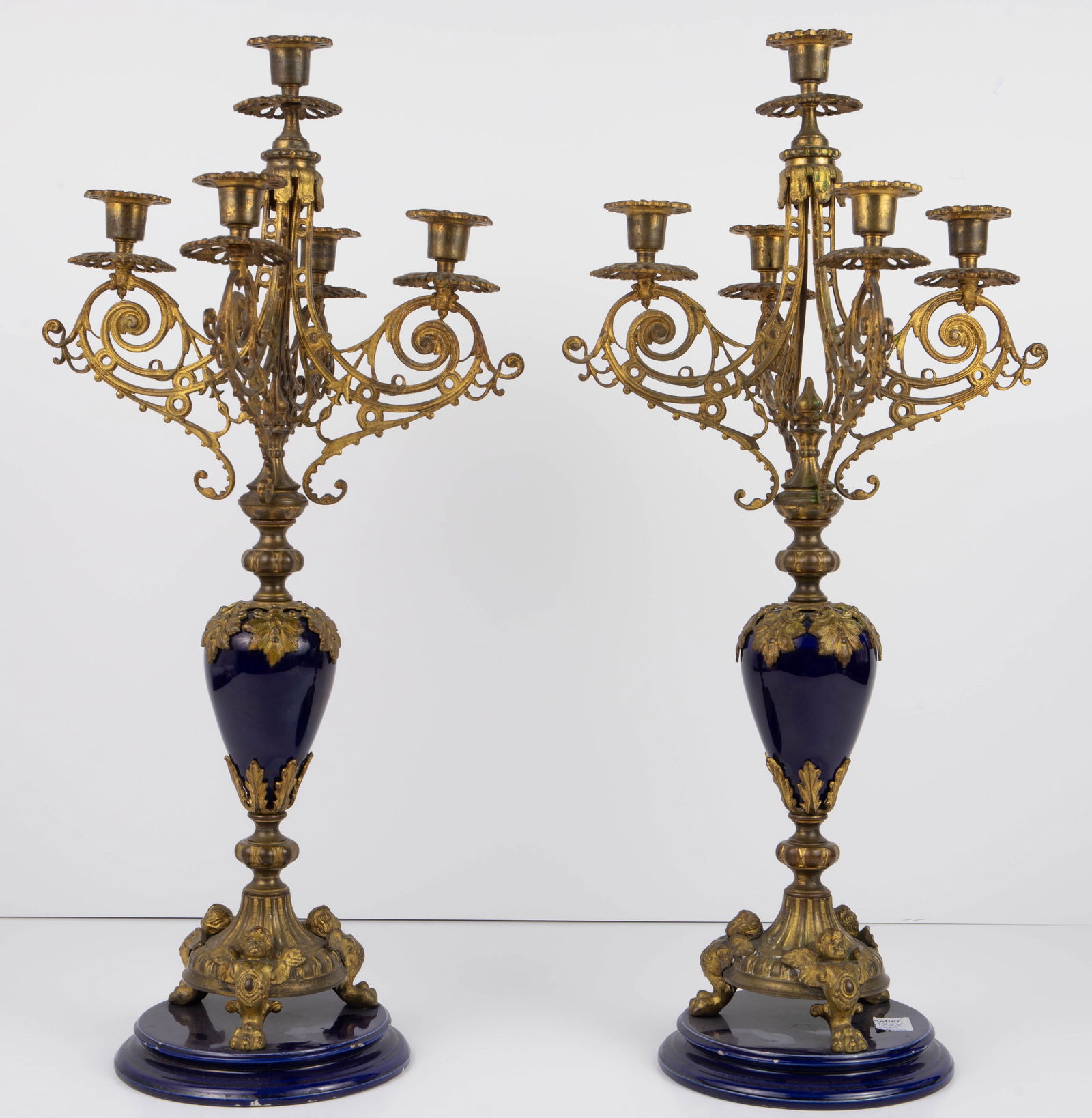 Pair of Bronze and Porcelain Cherub Candelabra.: These candelabra may be French; the maker is unknown. They are gilded bronze on cobalt blue porcelain, with three winged cherubs leading into paw feet, four scrolled arms with candle holders, and a ce