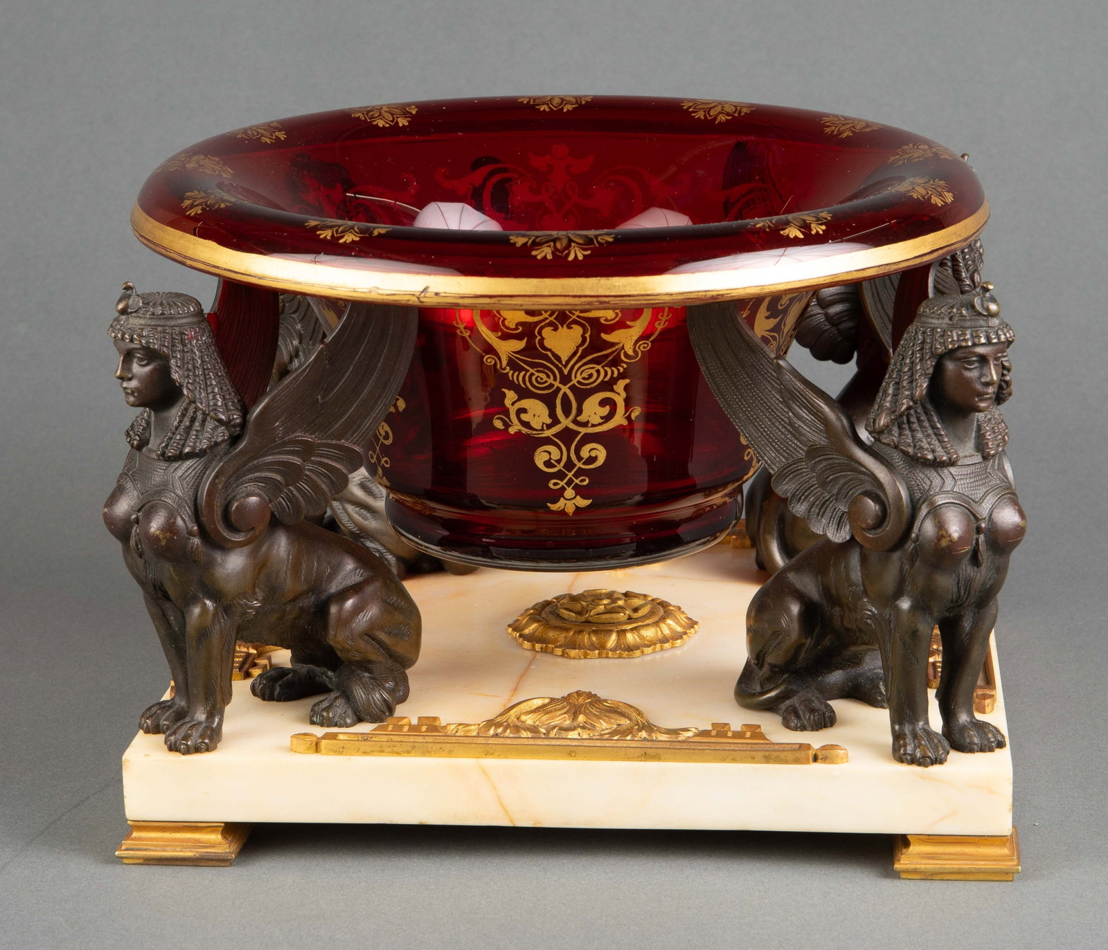 Marble, Bronze Sphinx & Cranberry Glass Bowl.: This cranberry glass and gilt bowl sits on four patinated bronze sphinx figurines, which sit on a cream marble slab. Also, the piece has ormolu bronze details and feet. The bowl does not quite fit on 