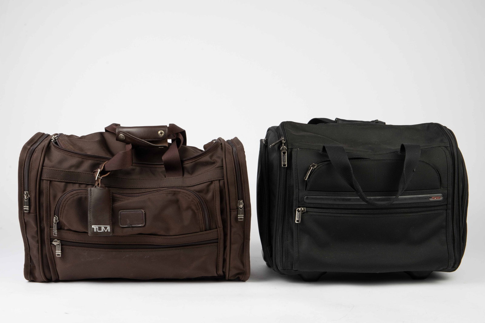 Pair Tumi Travel Bags - Garment, Satchel, Hangers.: The lot contains 2 Tumi carry-on travel bags. First, there is a black, 2 wheeled garment bag, which includes 6 Tumi hangers. Next, there is a brown satchel or duffel bag. The largest bag measures 14" 