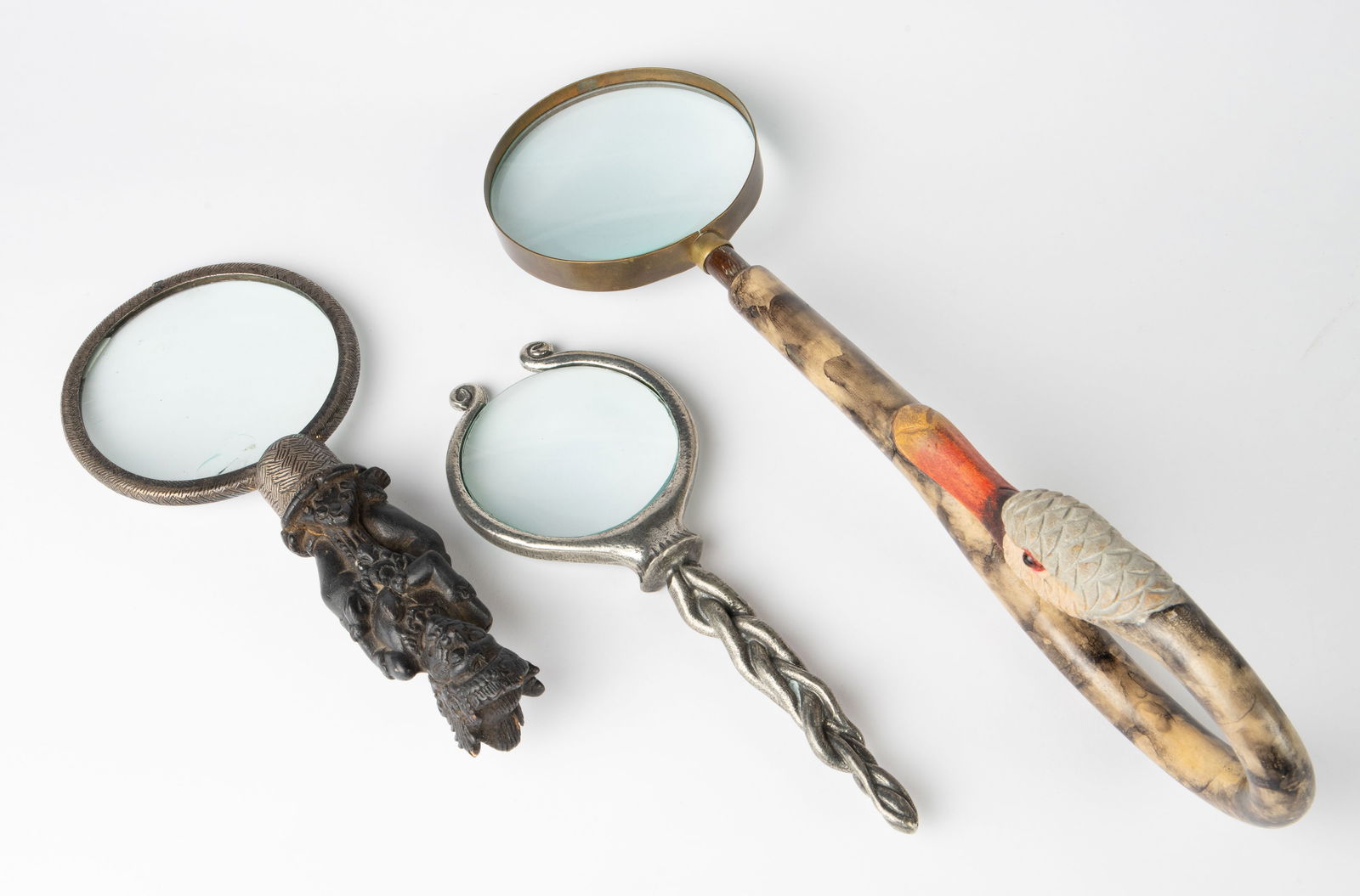 Three Magnifying Glasses - John Hardy, etc.: The lot contains 3 distinct magnifying glasses. First, there is a piece with a carved and painted wood handle depicting a duck. Next, there is a sterling silver and wood Indonesian piece with a carved