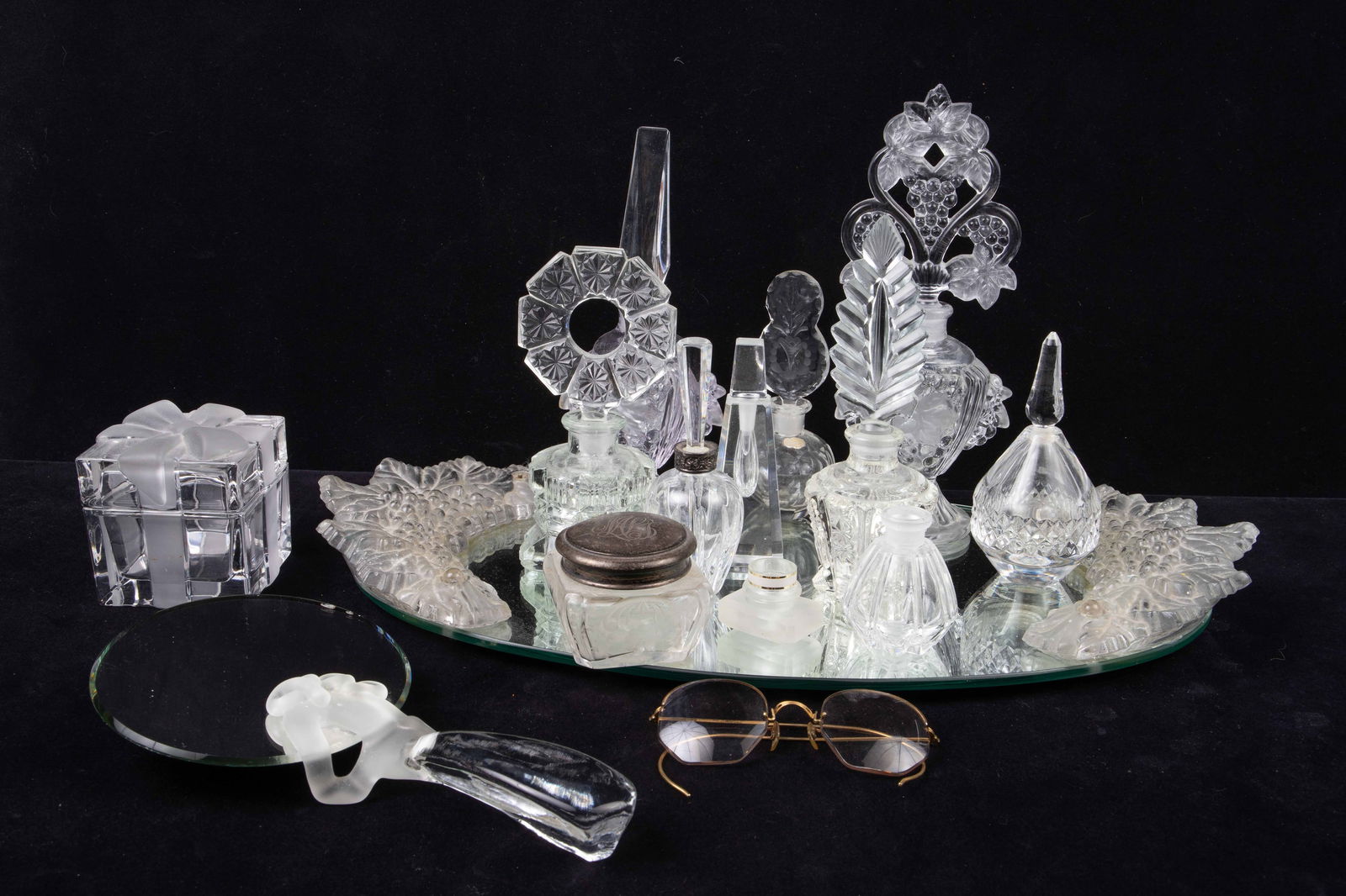 Lot of Glass Perfume Bottles w/ Vanity Tray.: This lot includes several glass and crystal perfume bottles, some signed. Also included are a signed hand mirror, a present-shaped trinket box, a pair of 10K gold filled eyeglasses, a vanity jar with