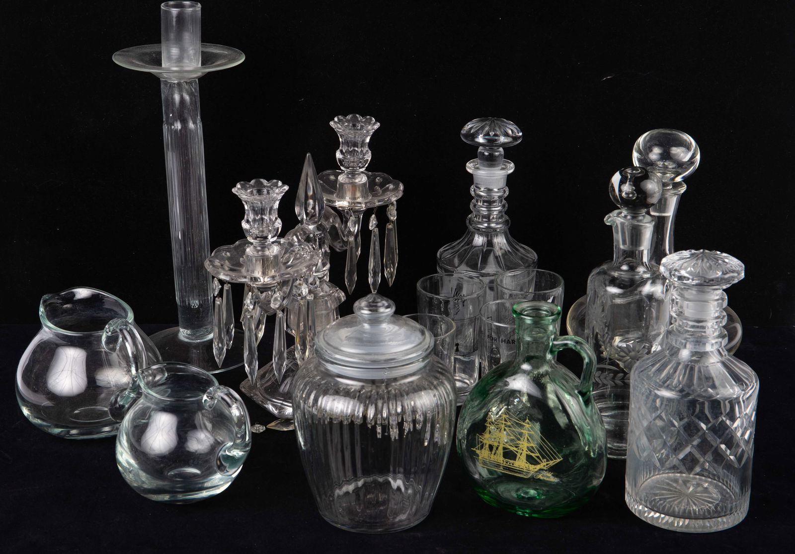 Lot of Glass Decanters, Jugs, Candle Sticks, Etc.: The lot contains 14 items in total. There are 5 decanters (4 with stoppers), a lidded jar, 2 jugs, 4 tumblers, a 2 arm candelabra with prisms, and a single large candlestick. The green glass decanter,