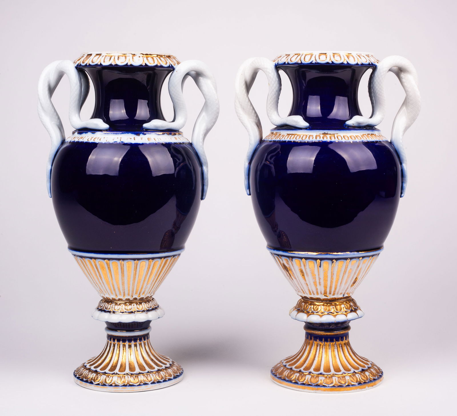 Pair of Meissen Amphora Vases w/ Serpent Handles.: These cobalt blue, gold, and white snake handle vases each bear a blue crossed swords mark underneath, along with the handwritten alphanumeric code, "E153". Each vase or urn has some gilt loss. One of