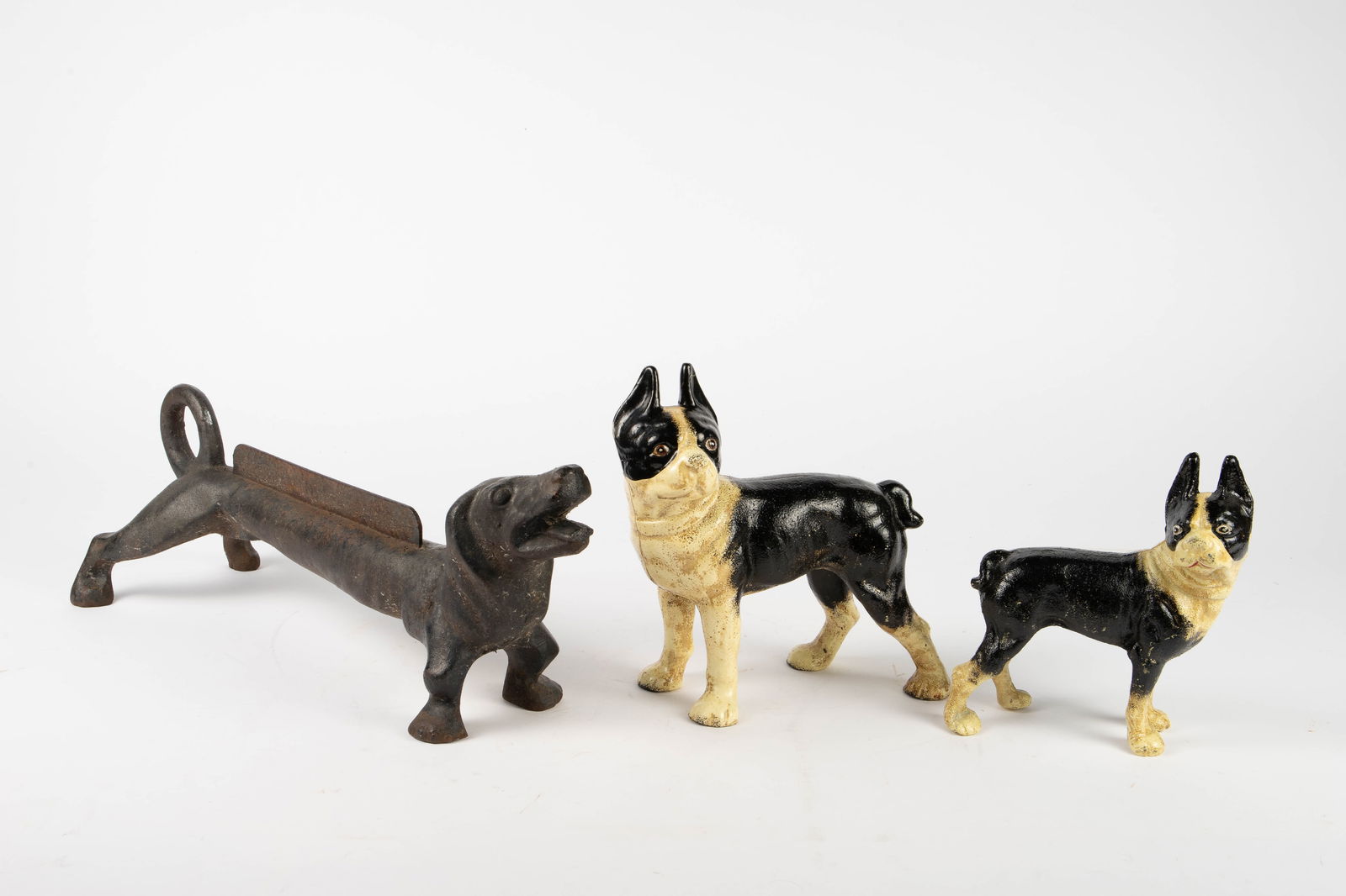 Lot of 3 Cast Iron Dogs - Doorstops, Boot Scraper.: The boot scraper resembles a very long dachshund. The other two dogs are black and white Boston Terriers. Overall, the lot looks to be in good condition, with some surface wear. The largest item measu